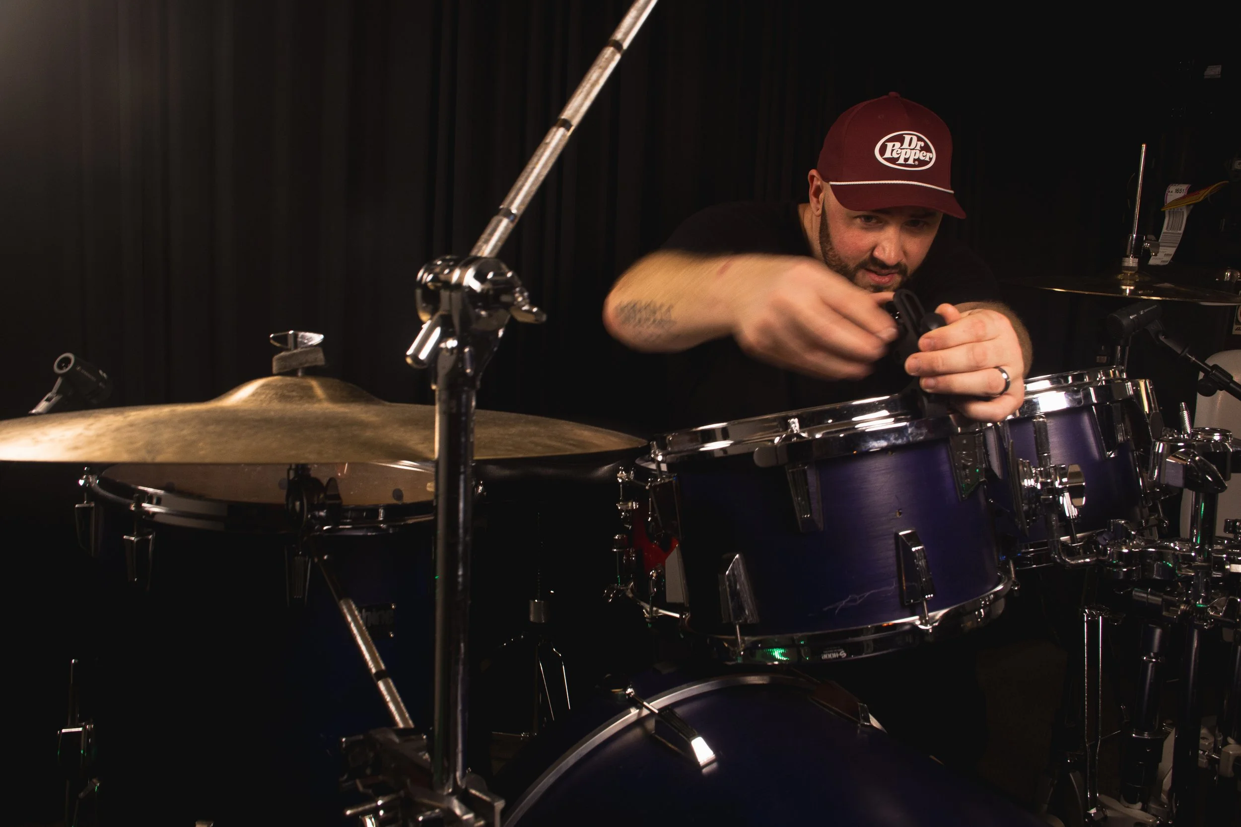Drummer Greg Vance of the band Syr, wearing a maroon baseball cap with a 'Dr. Pepper' logo and a black shirt is adjusting a drum microphone at Dreamscape Co-op in Irmo, SC