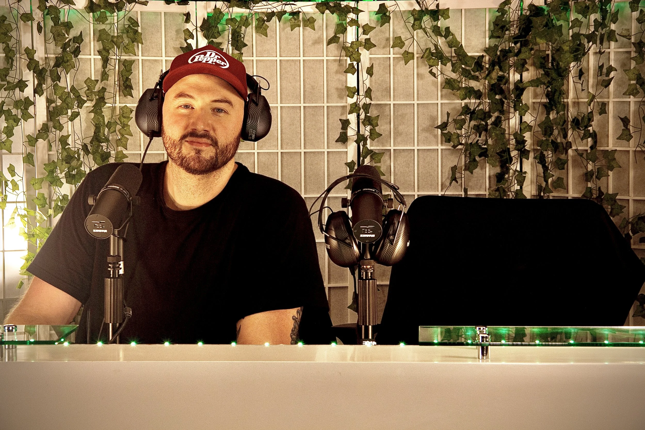 A man sitting at a desk with a microphone and headphones, in front of a wall decorated with artificial green ivy and string lights.