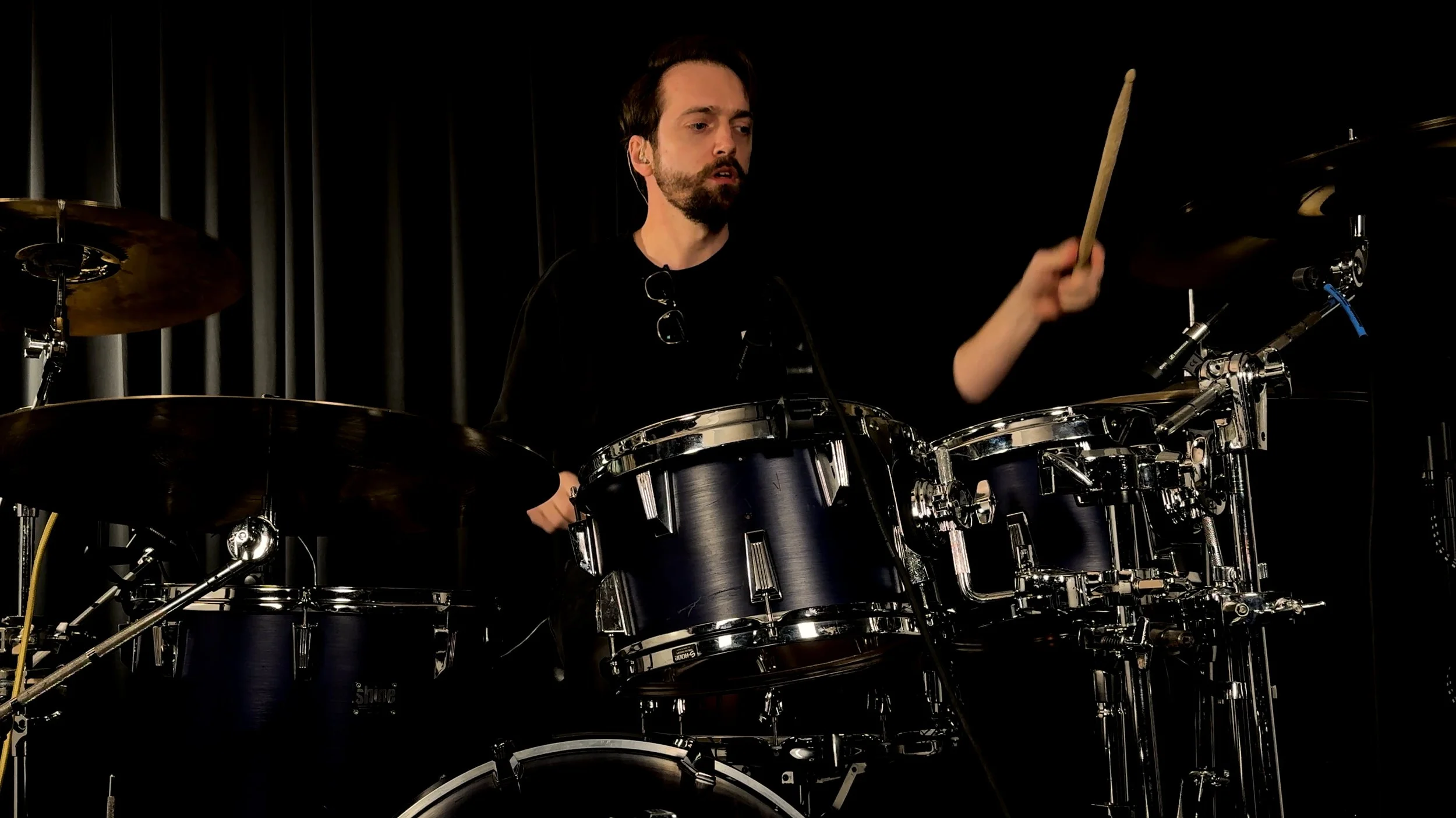 A man playing a drum set on stage, with a dark background and curtain.