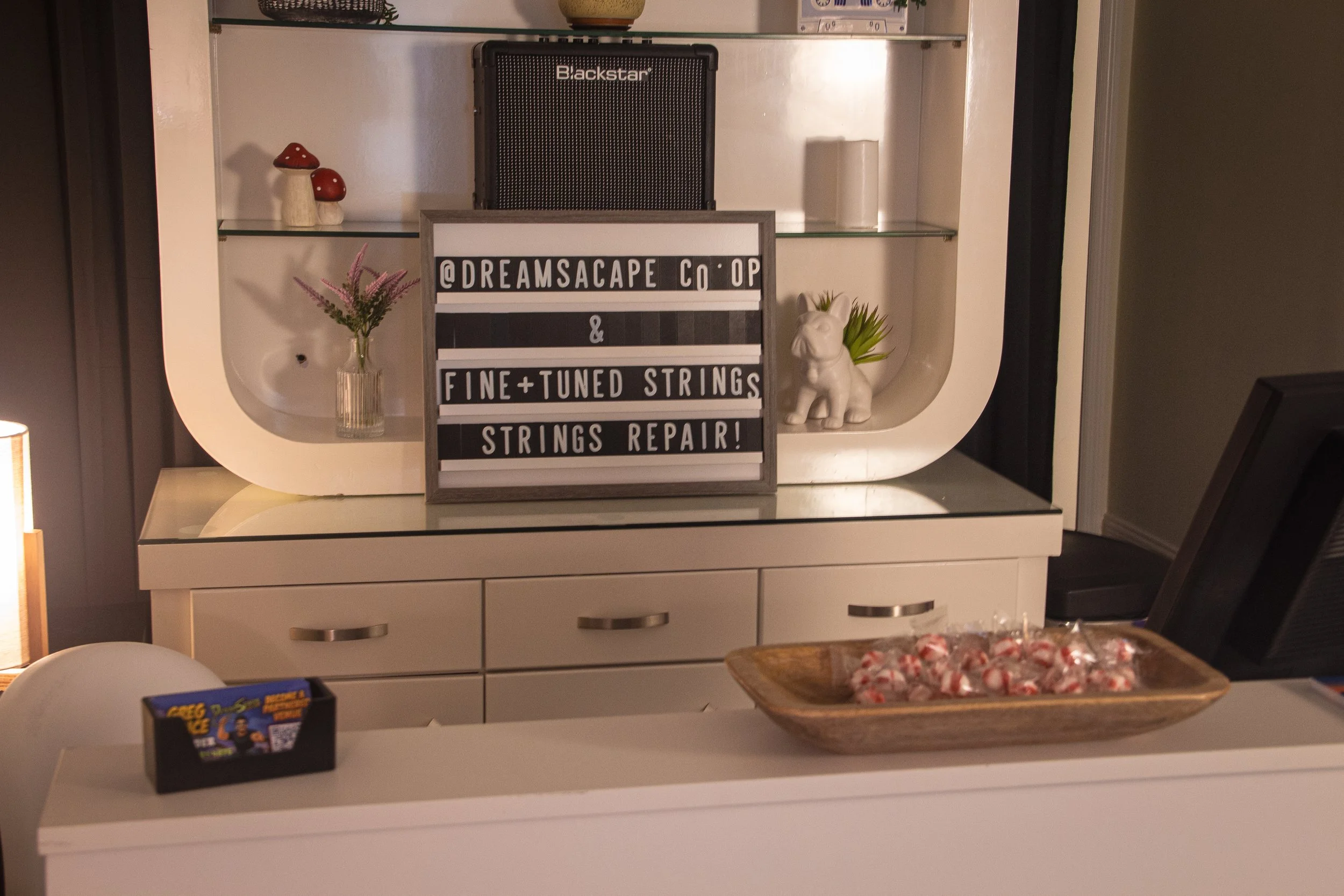 Decorative display in a cabinet with a sign that reads '@DREAMSACAPE Co-OP & FINE-TUNED STRINGS STRINGS REPAIR!', small mushroom figurines, a potted plant with flowers, a small elephant statue with plants behind it, a black speaker, and a wooden tray of candies on a white surface.