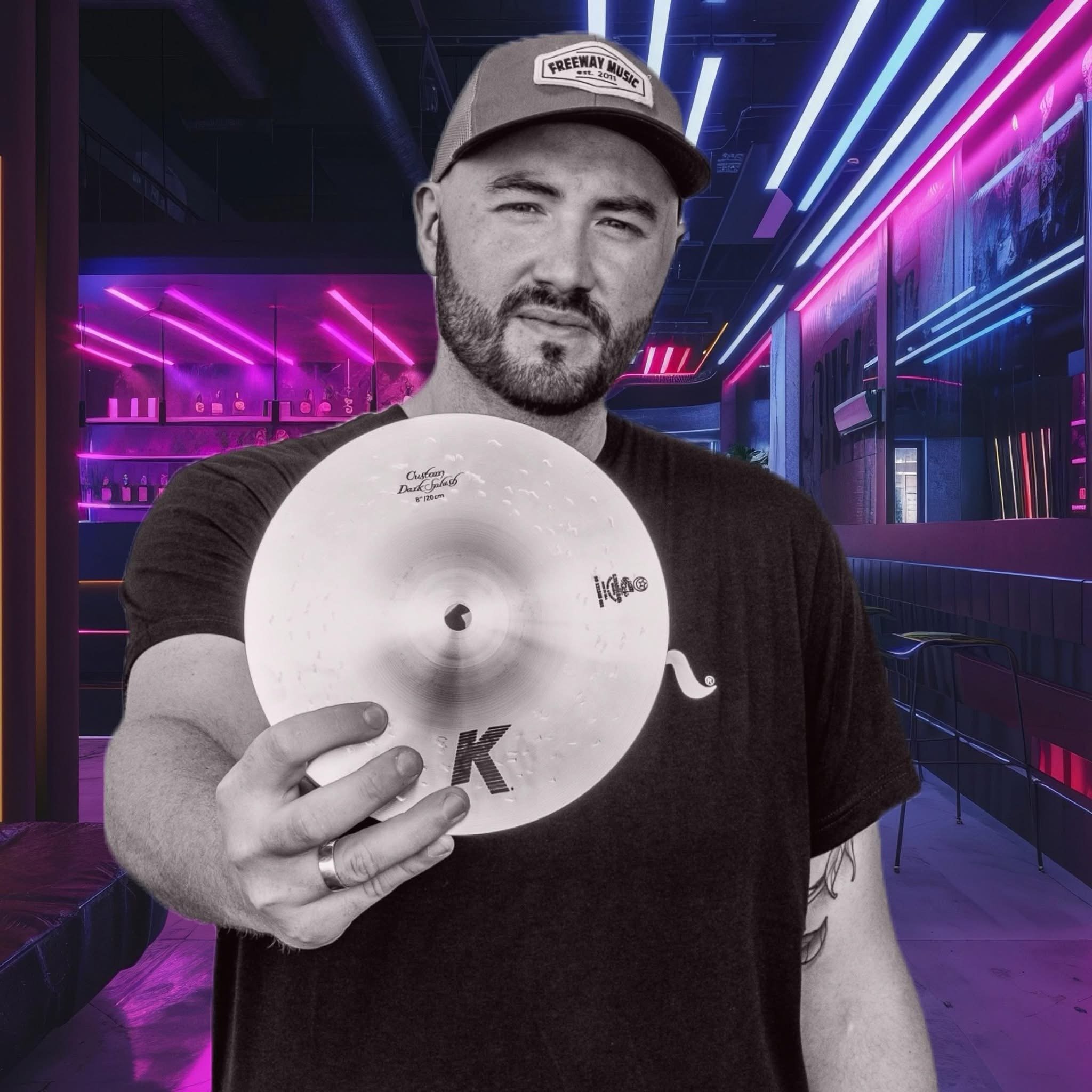 Drummer Greg Vance with a beard wearing a baseball cap holding a Zildjian K splash cymbal, in a neon-lit bar or nightclub with purple and pink lighting. Advertising his Social Media, Podcast, and content studio in Columbia, SC