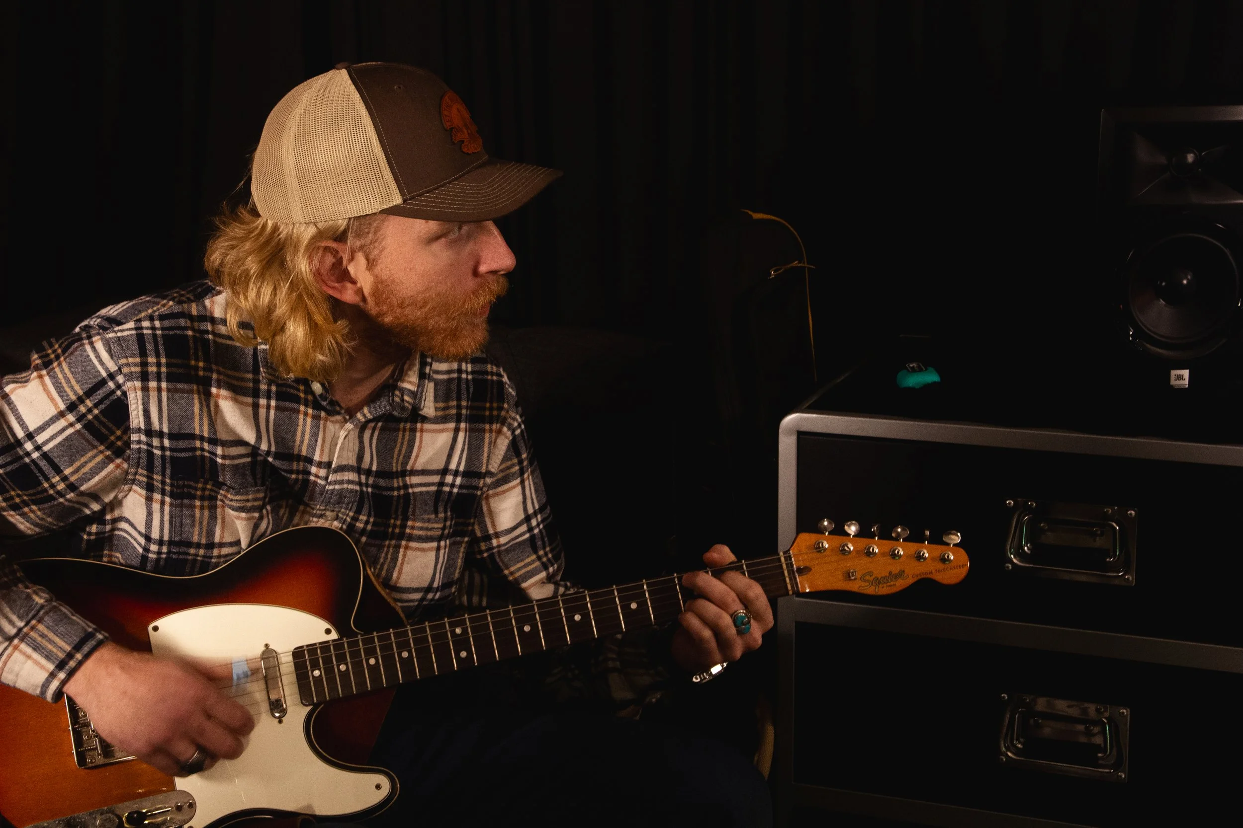 A man with long blonde hair and a beard wearing a plaid shirt and a trucker hat, playing an electric guitar in a dimly lit room.