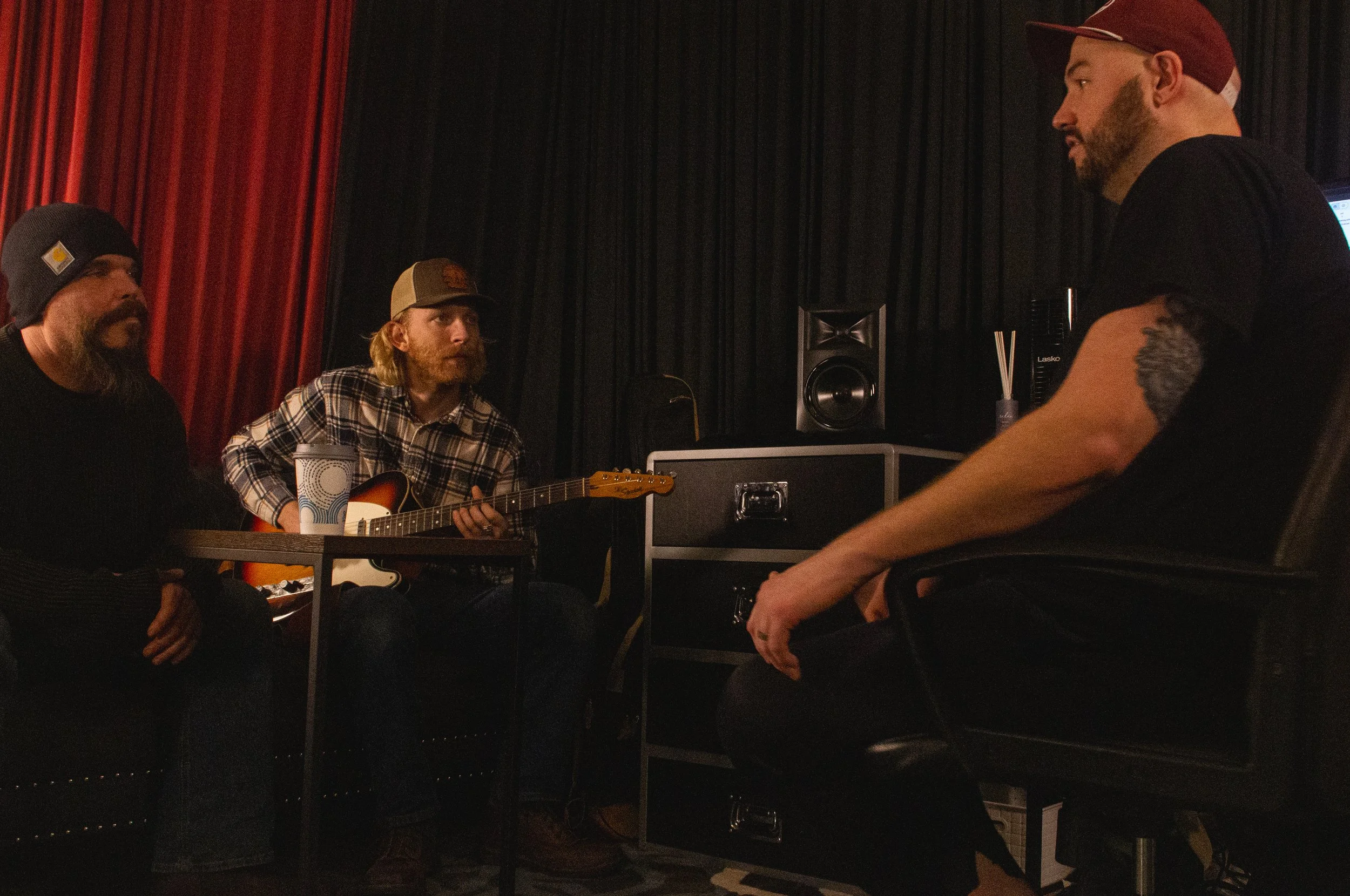 Drummer Greg Vance, Drayton Brookes, and Dale Kennedy in the Dreamscape Co-op recording studio; two sitting on a couch, one facing them with a guitar, engaged in conversation. Photo by Philip Thomas
