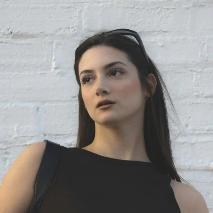 A young woman with long dark hair, wearing a black sleeveless top and sunglasses on her head, stands in front of a white brick wall.