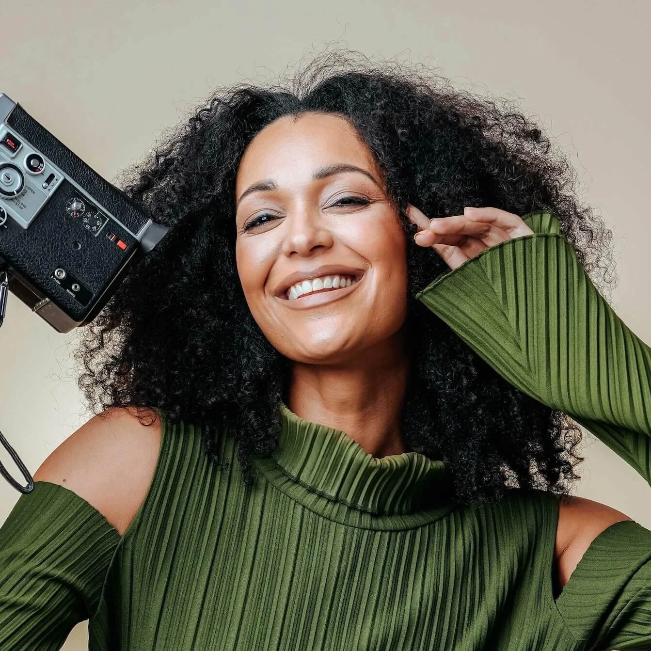 A woman with curly black hair is smiling and posing for a photo in front of a camera.