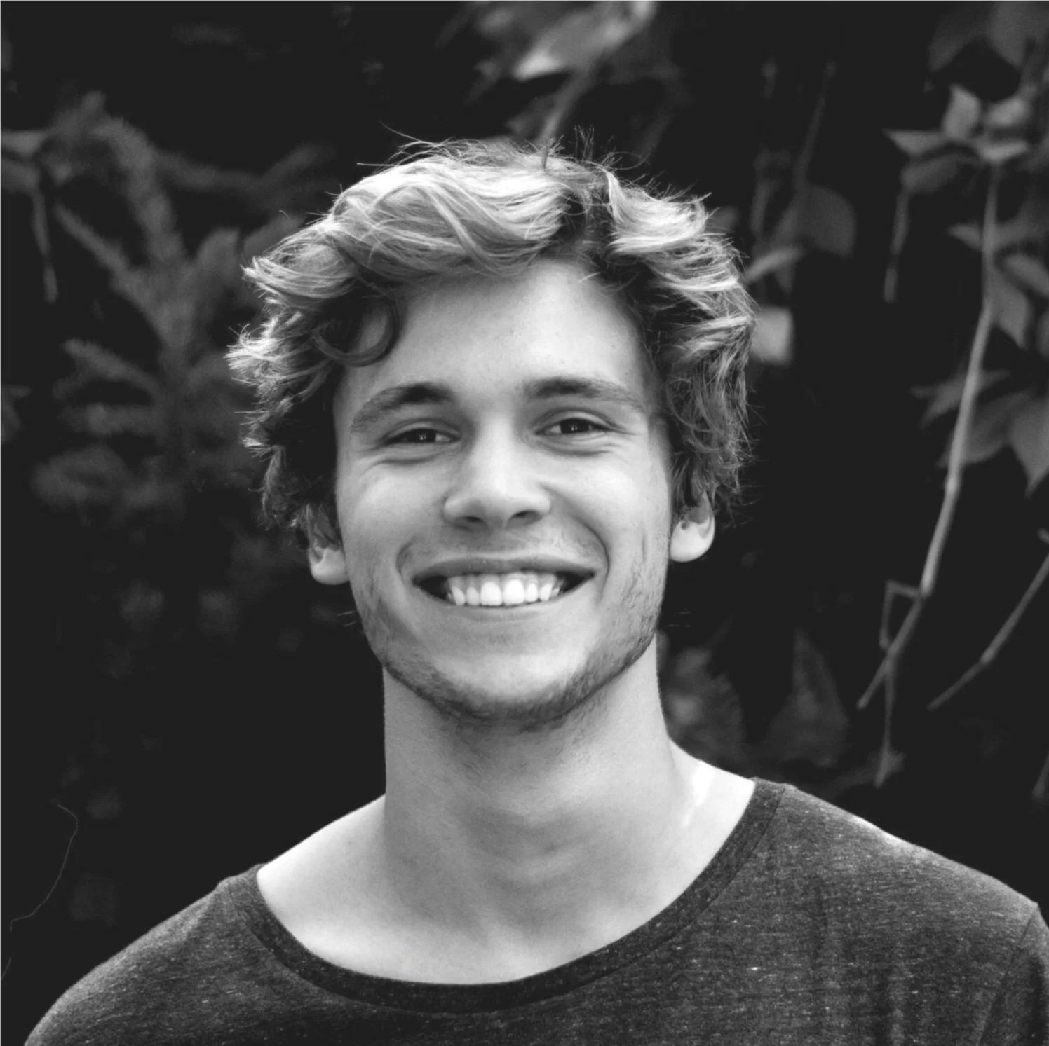 Black and white photo of a young man with curly hair, smiling, wearing a dark T-shirt, standing outdoors with blurred foliage in the background.