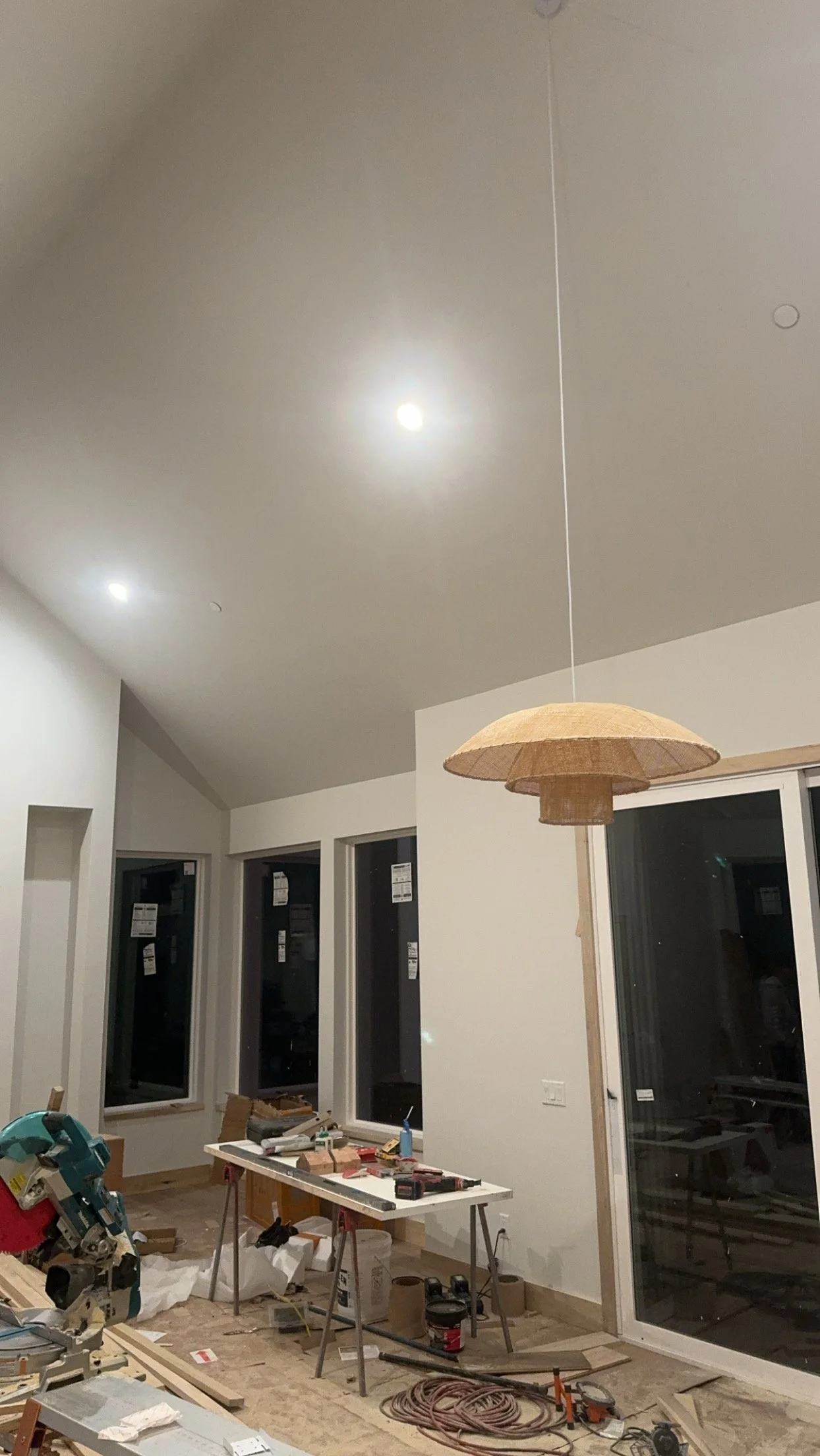 A room under construction or renovation, with a white table holding tools, and a ceiling light fixture hanging from the ceiling. The room has large windows and a sliding door.