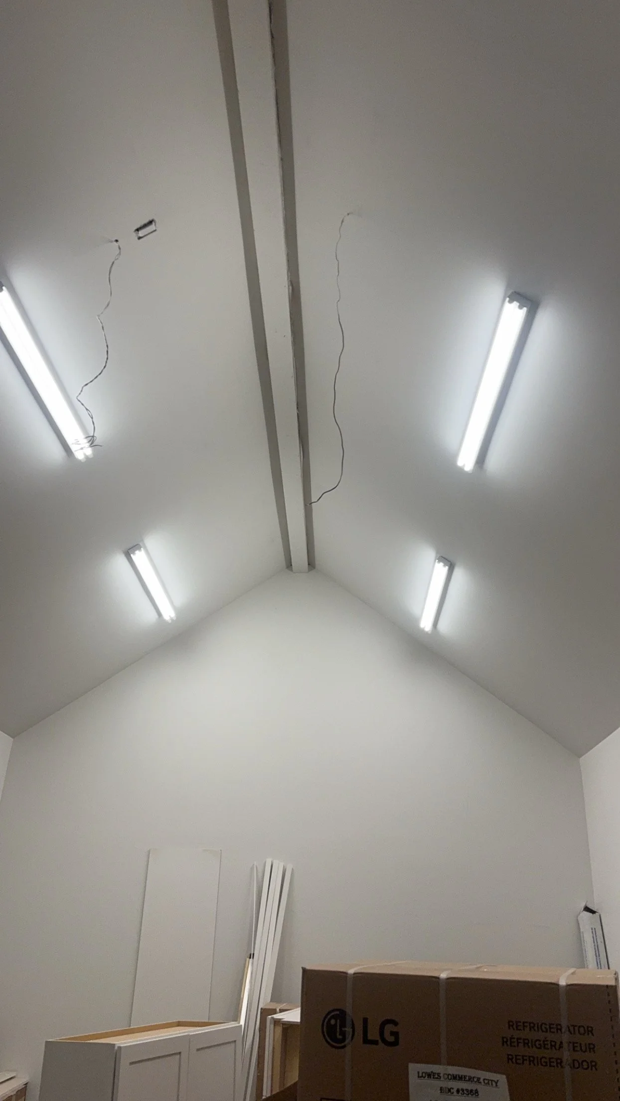 Ceiling of a room with four fluorescent light fixtures, connecting wires hanging from the ceiling, and a few boxes and furniture at the bottom.