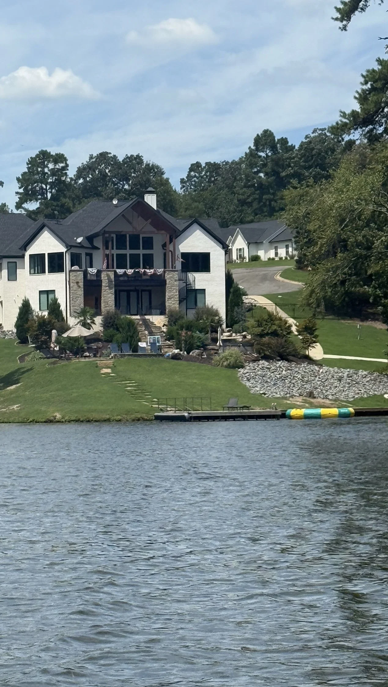 A large modern house with white walls, dark roof, and large windows by a body of water. The house has a back patio with umbrellas and outdoor furniture, surrounded by greenery and rocks.