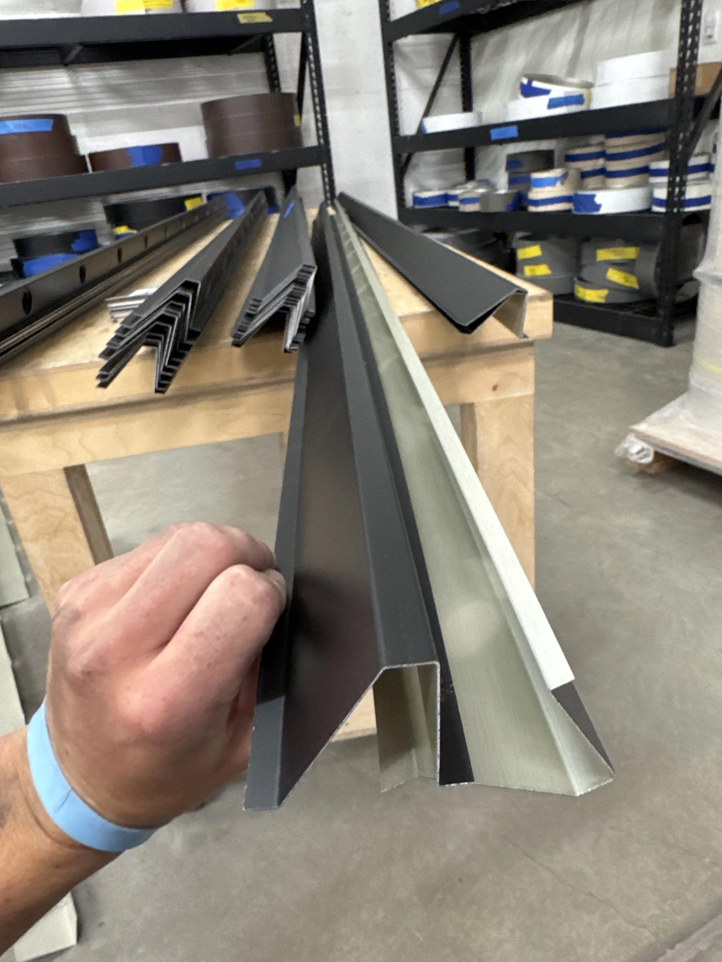 Close-up of a person's hand holding a metal framing stud in a hardware store with shelves of various metal pieces and tapes in the background.