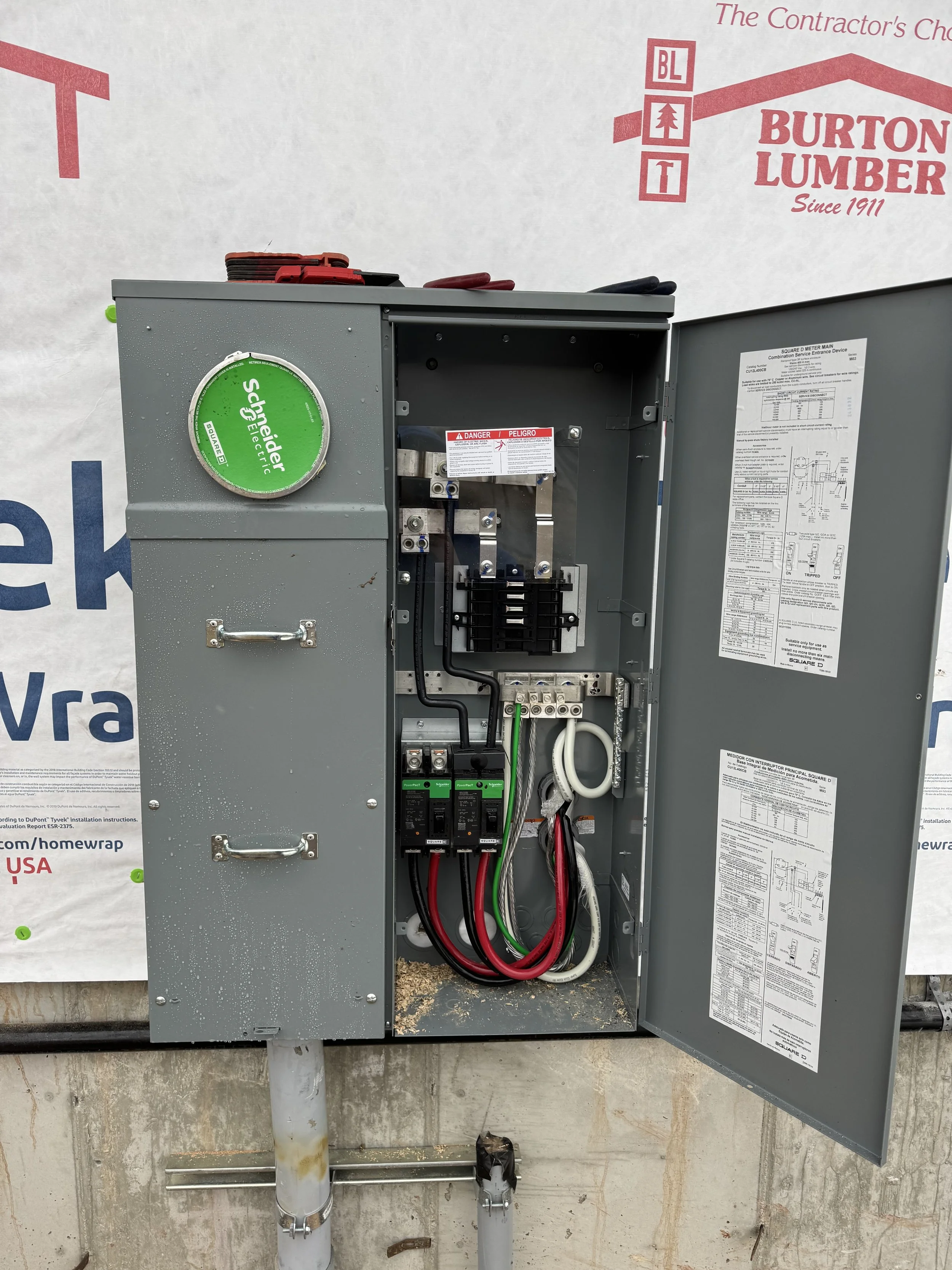 Electrical panel box with wiring, circuit breakers, and safety labels, mounted on a post outside next to a wall with construction company branding.