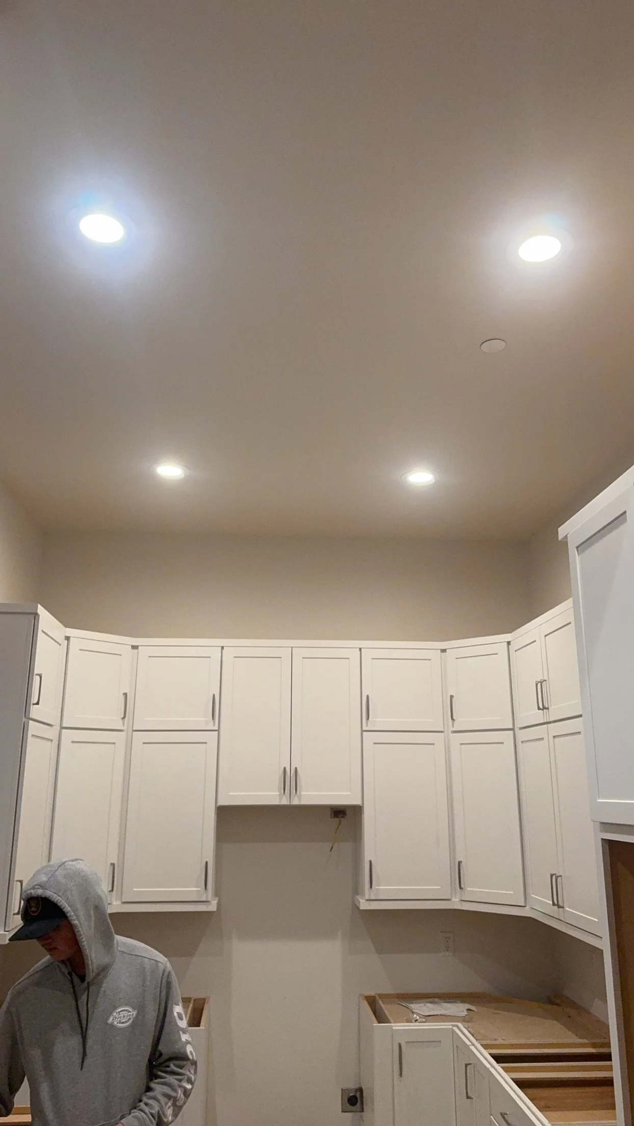 A person working in a kitchen with white cabinets and a beige wall. The ceiling has four recessed lights.