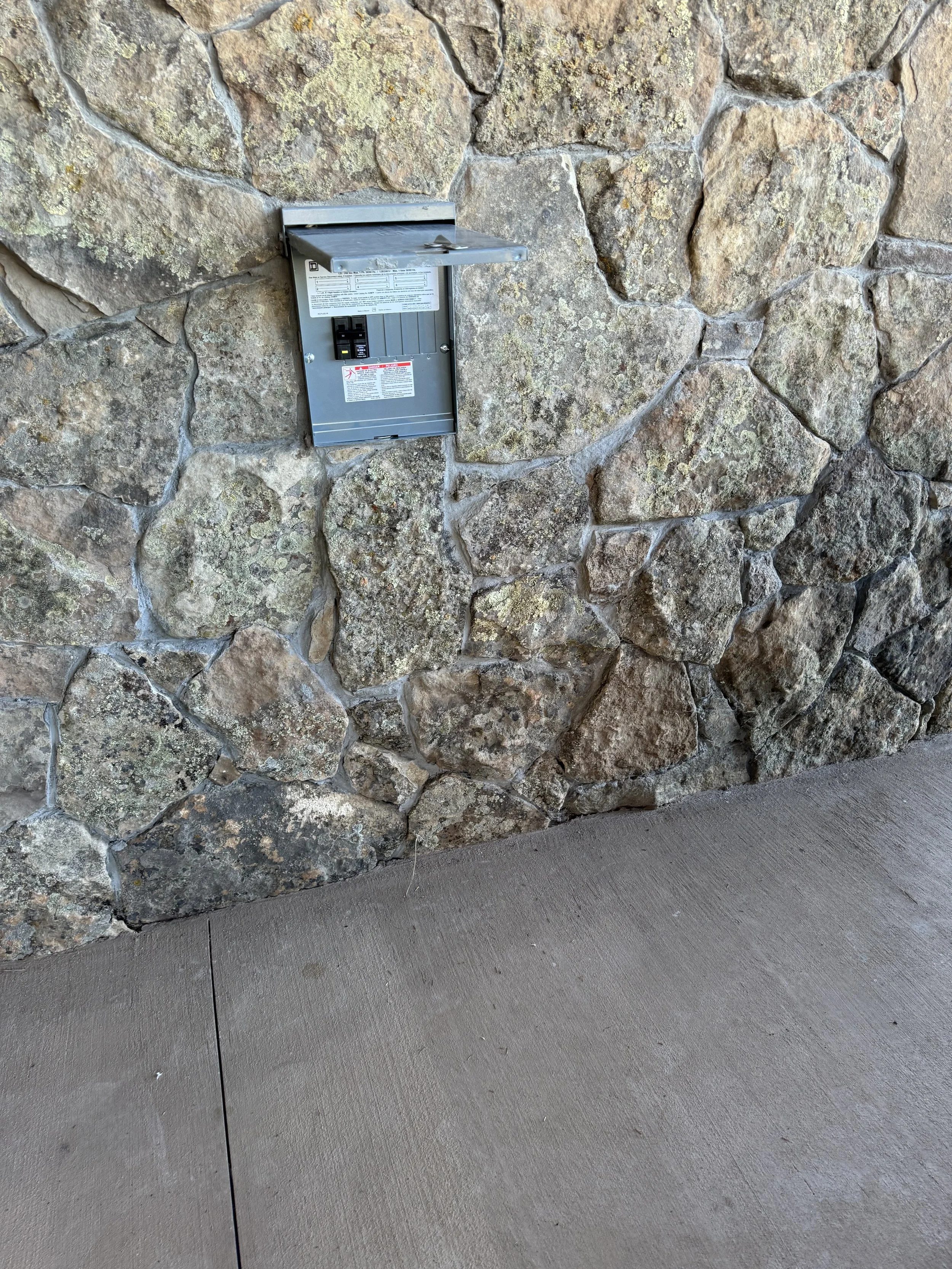 Electric meter box mounted in a stone wall with a concrete sidewalk in front.