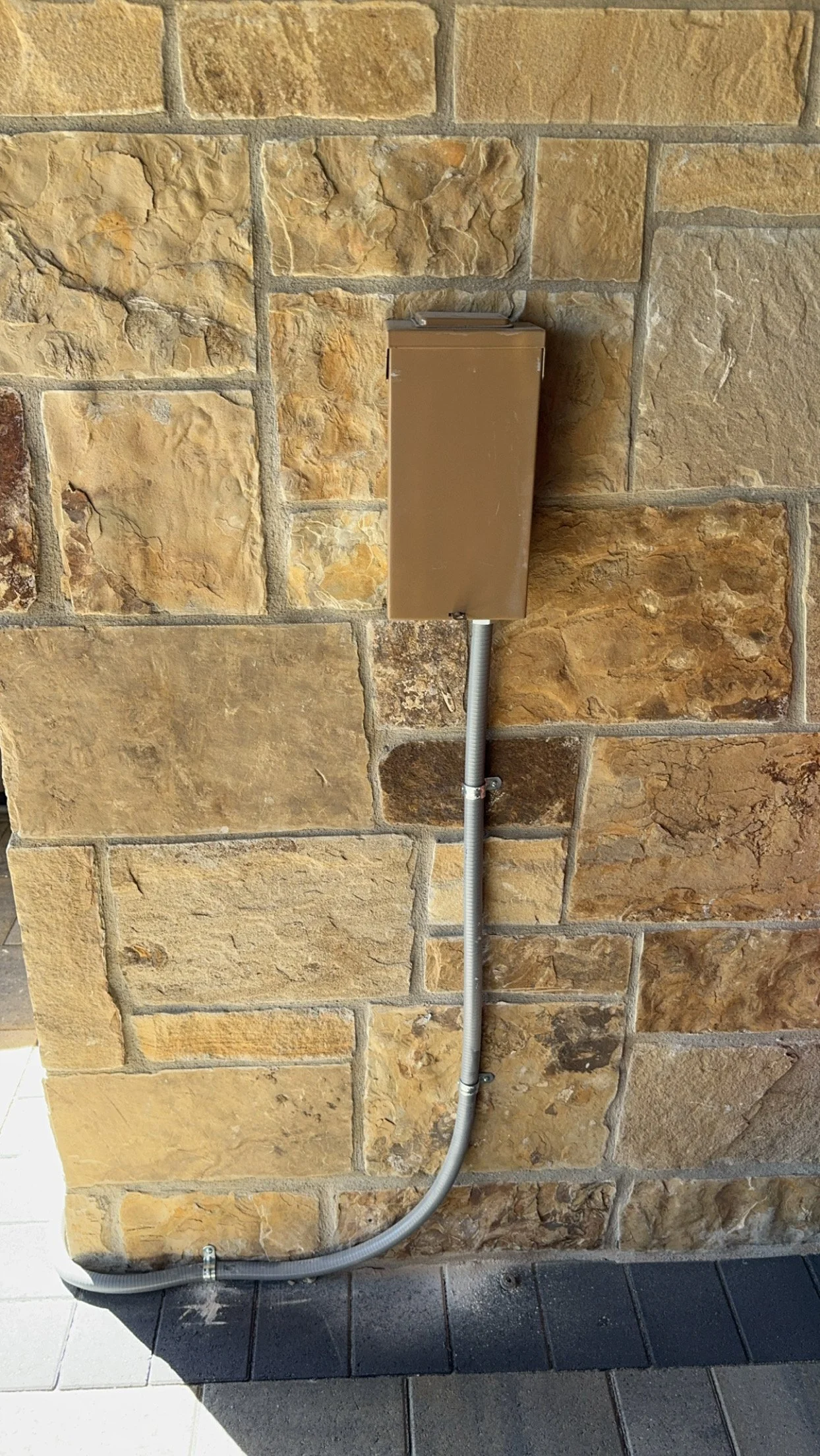A beige electrical box mounted on a stone wall with an electrical conduit running down to the ground.