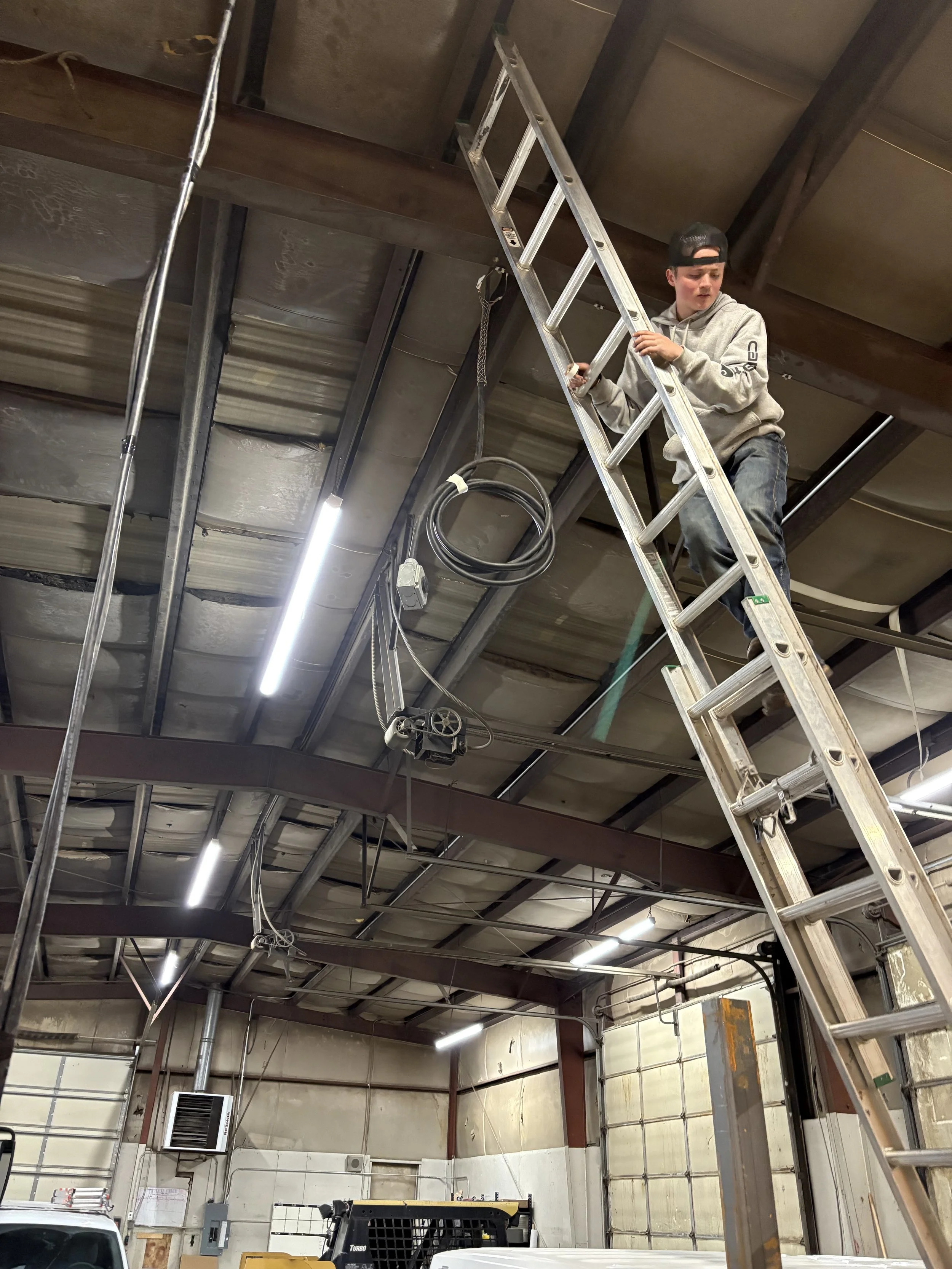 A young man in casual clothes and a cap is standing on a tall ladder, working on the ceiling of an industrial building with exposed metal beams, wires, and insulation.