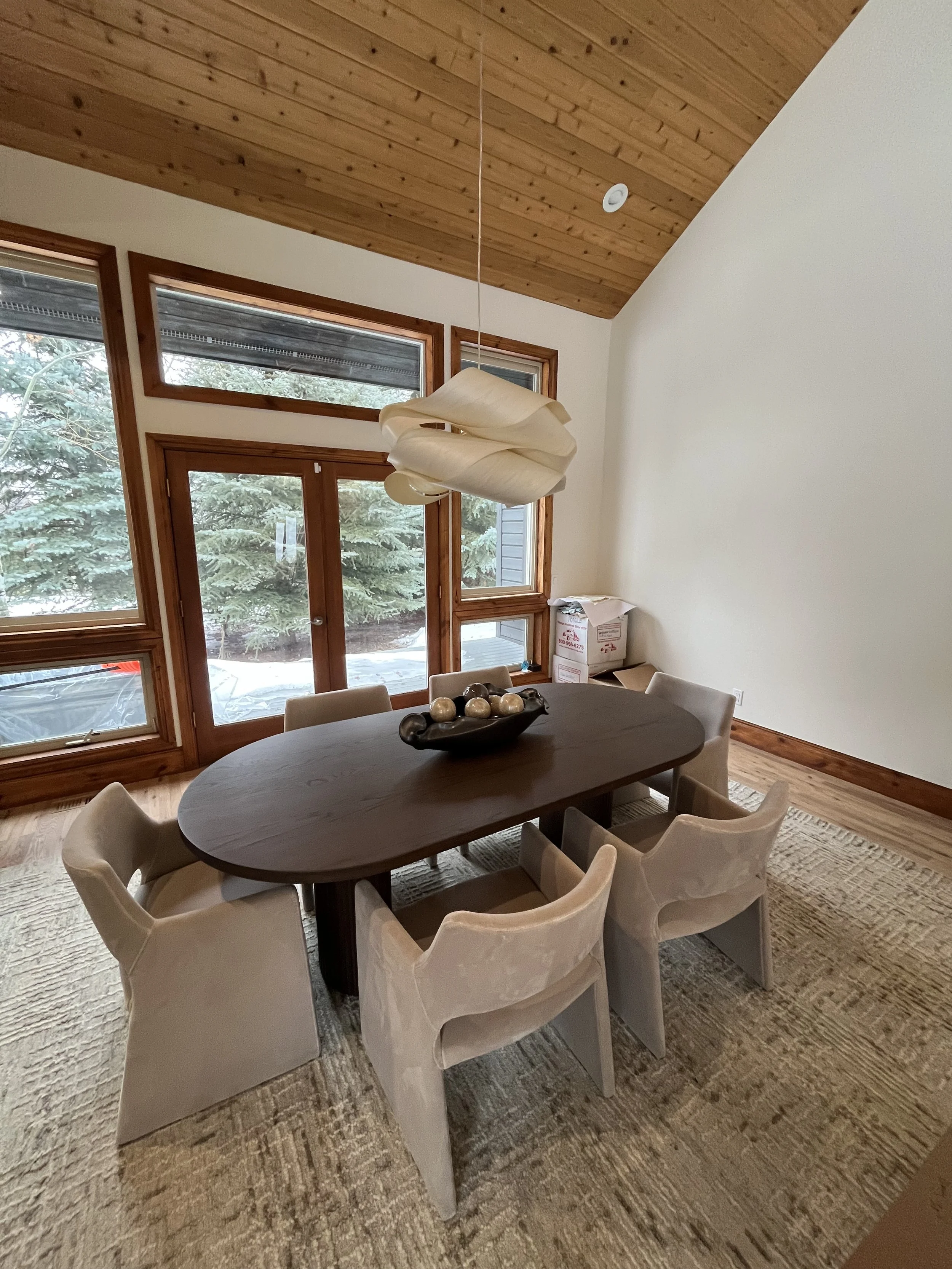 Dining room with a wooden table surrounded by six beige chairs, large windows with wooden frames, a wooden ceiling, and a modern hanging light fixture, with cardboard boxes in the corner.