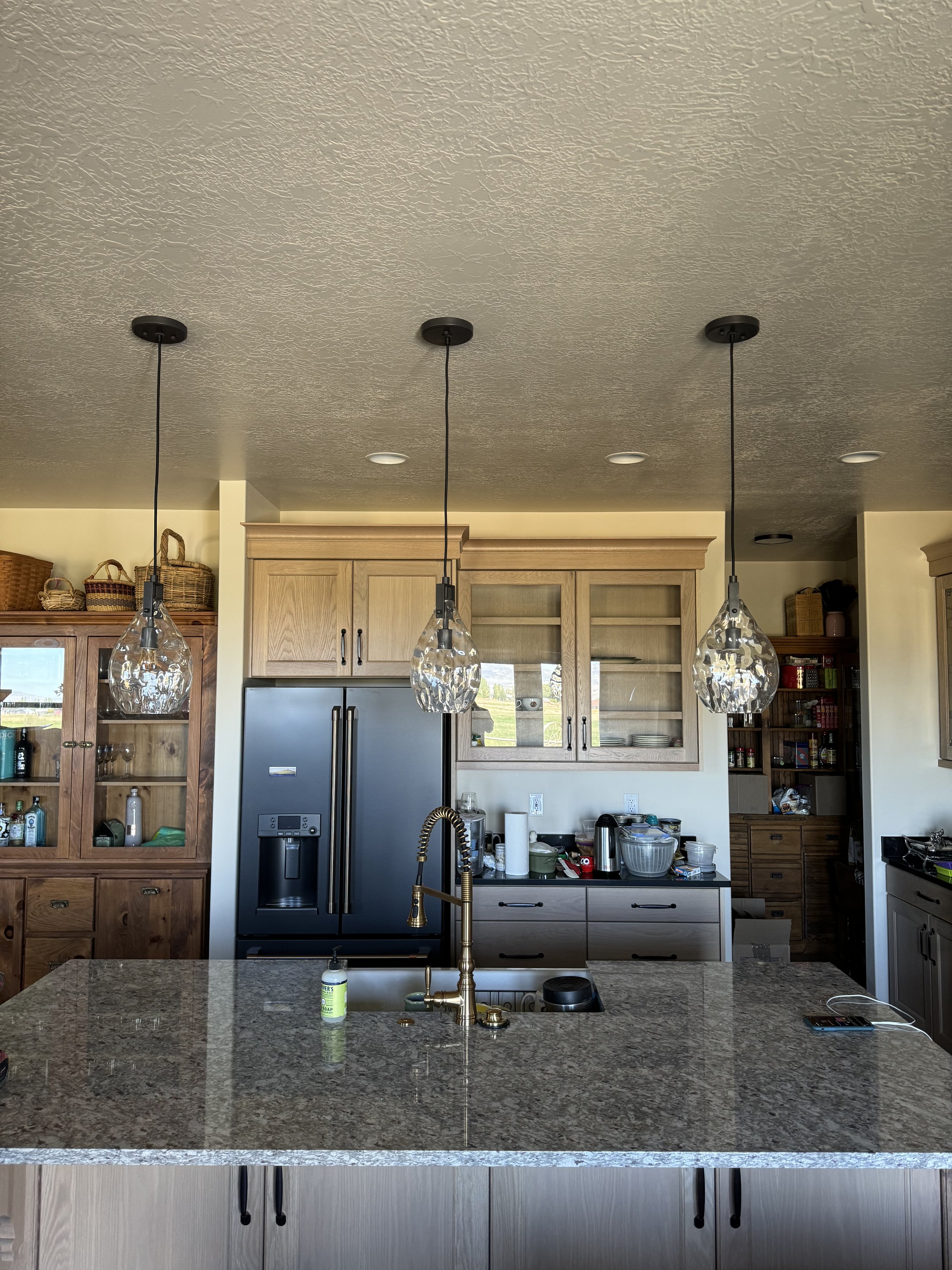 A modern kitchen with a granite island, pendant lights, wooden cabinetry, a stainless steel refrigerator, and various kitchen items on the counter