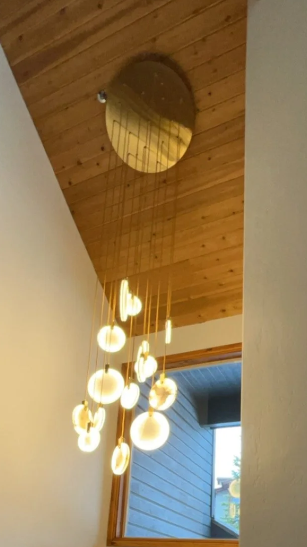 Modern hanging chandelier with multiple glowing round bulbs, installed in a room with wooden ceiling and trim, and a window showing trees outside.