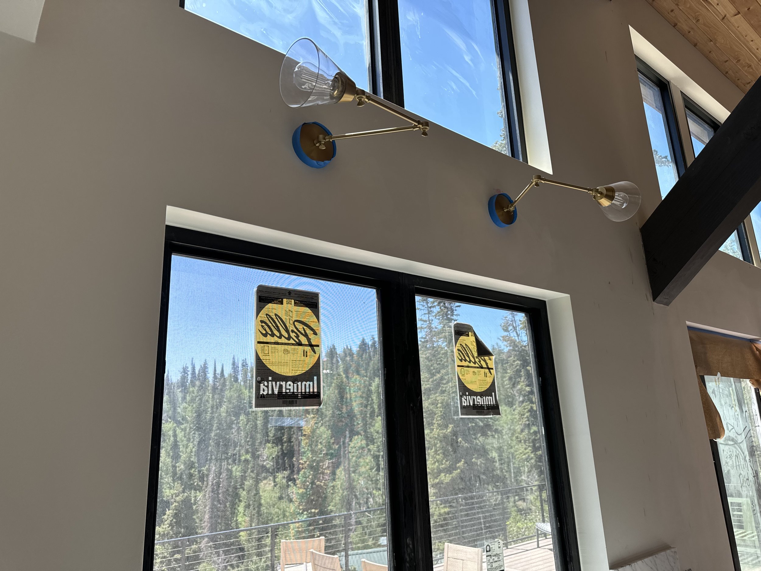 Modern interior with two brass and glass wall-mounted lamps above large windows, showing a view of trees and a blue sky.