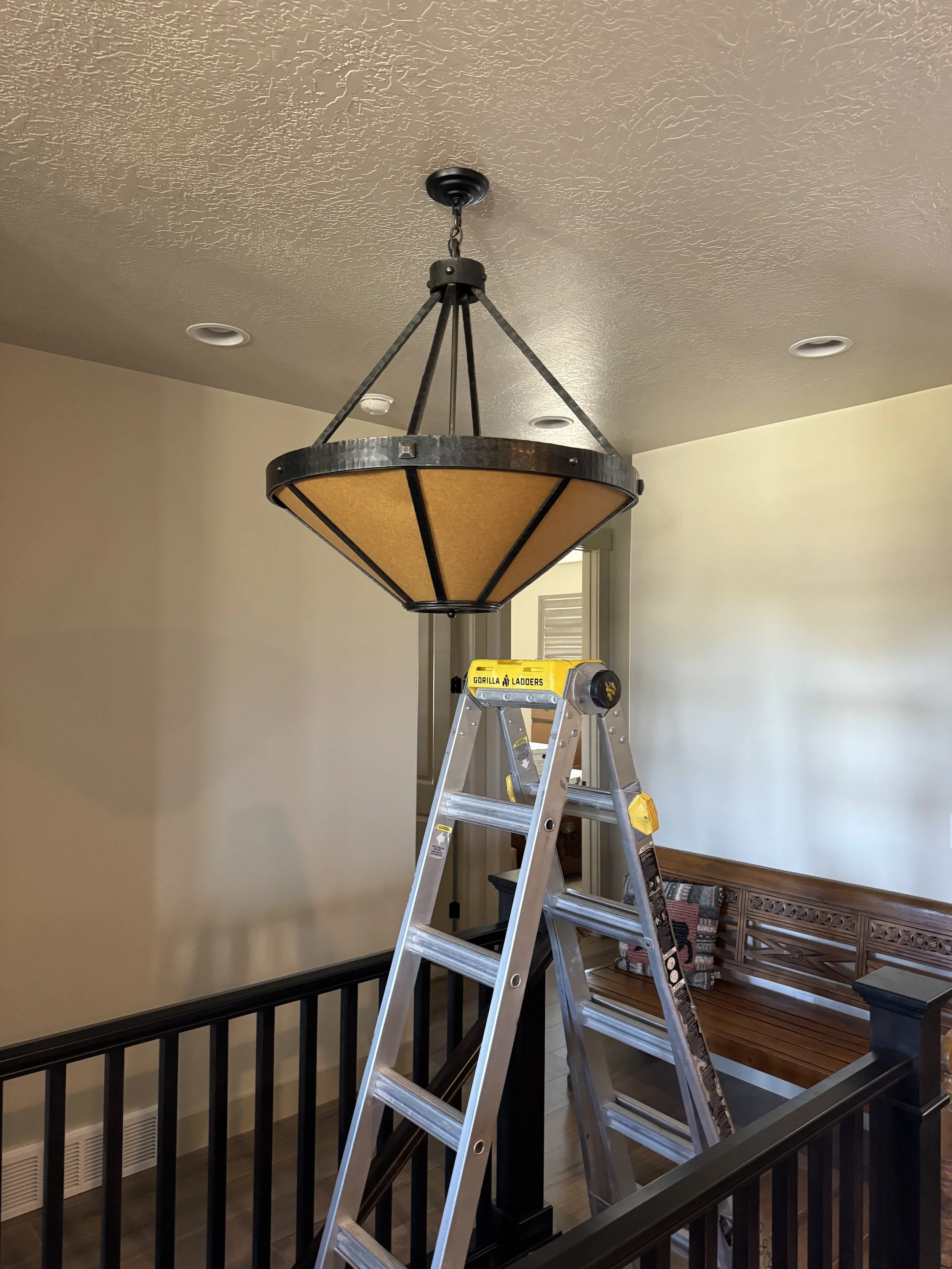 A ladder positioned underneath a ceiling light fixture, which is partly disassembled, in an indoor room with beige walls and a wooden bench.