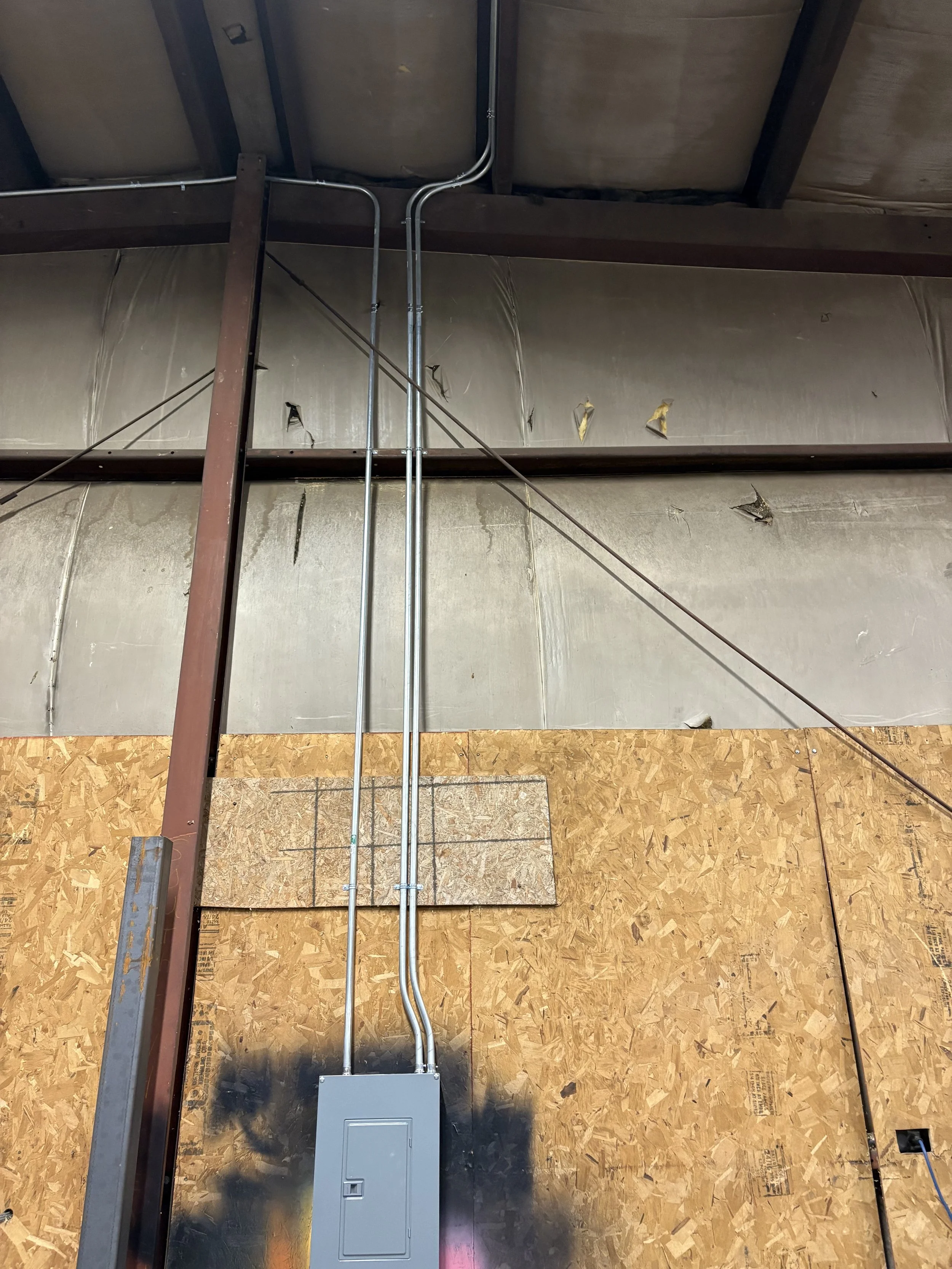 Electrical conduit pipes running down a furnace wall with a gray electrical box at the bottom, in an unfinished construction interior with plywood and metal framing.