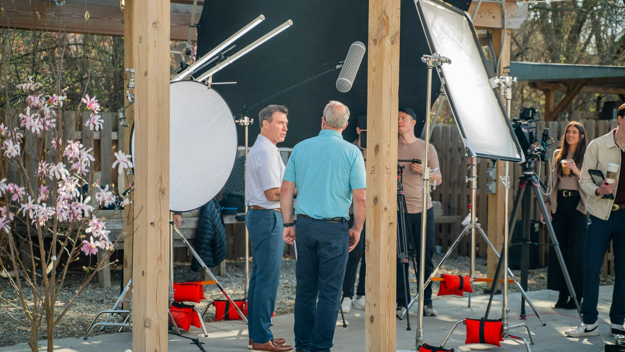 People filming a scene outdoors with professional video equipment, including lights and cameras, in a backyard setting.