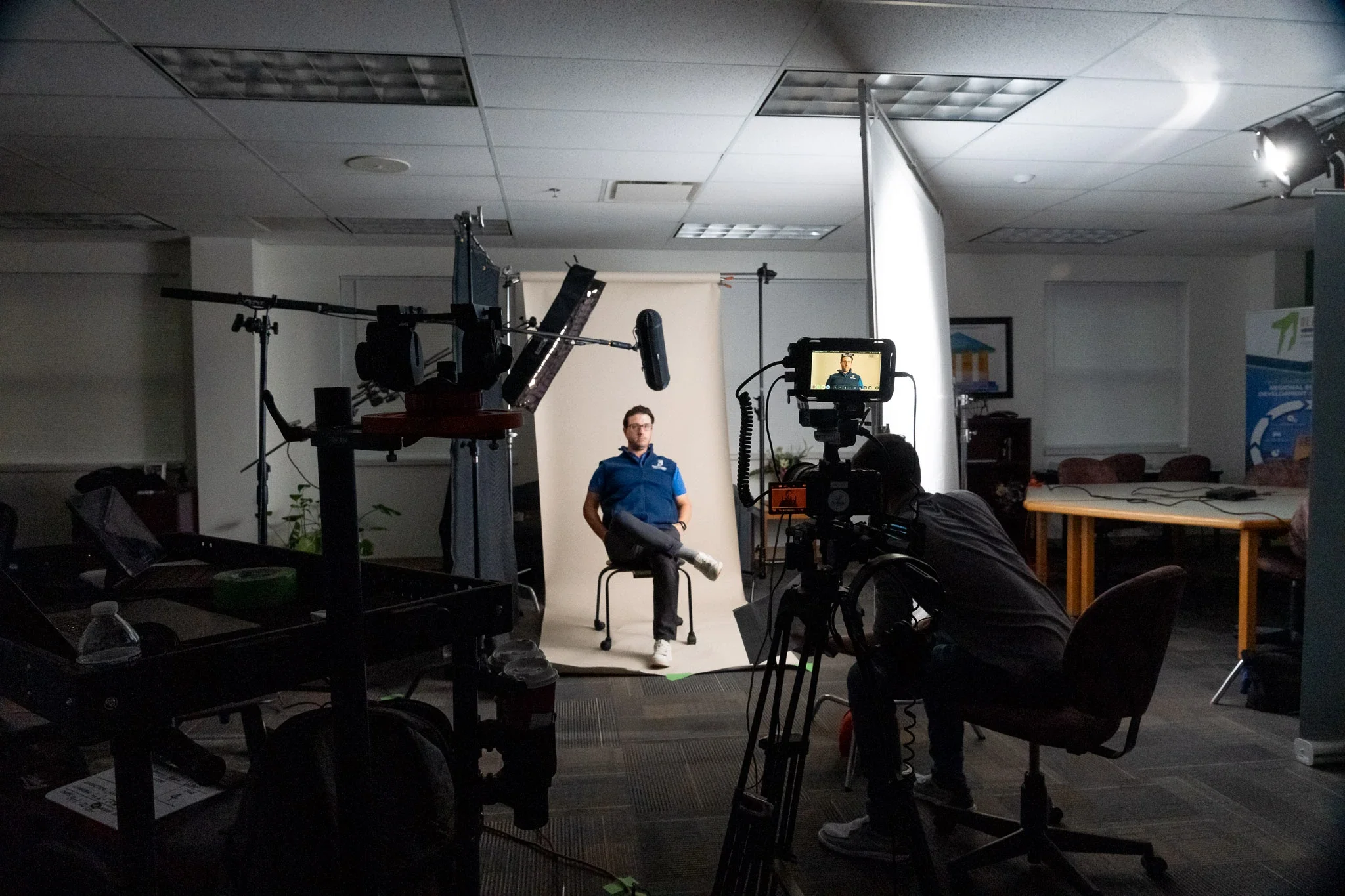 A man sitting on a chair in front of a camera and backdrop in a photography studio, with lighting and film equipment set up around him.