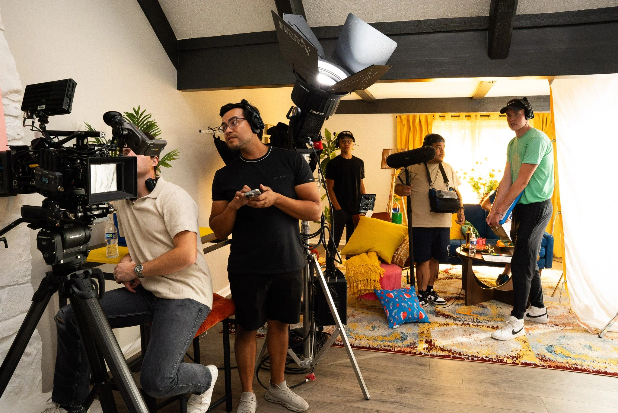A film crew is filming inside a living room with a colorful rug, yellow curtains, and a sofa with yellow and pink pillows. Several crew members are operating cameras and equipment, with one man sitting and others standing and holding boom microphones