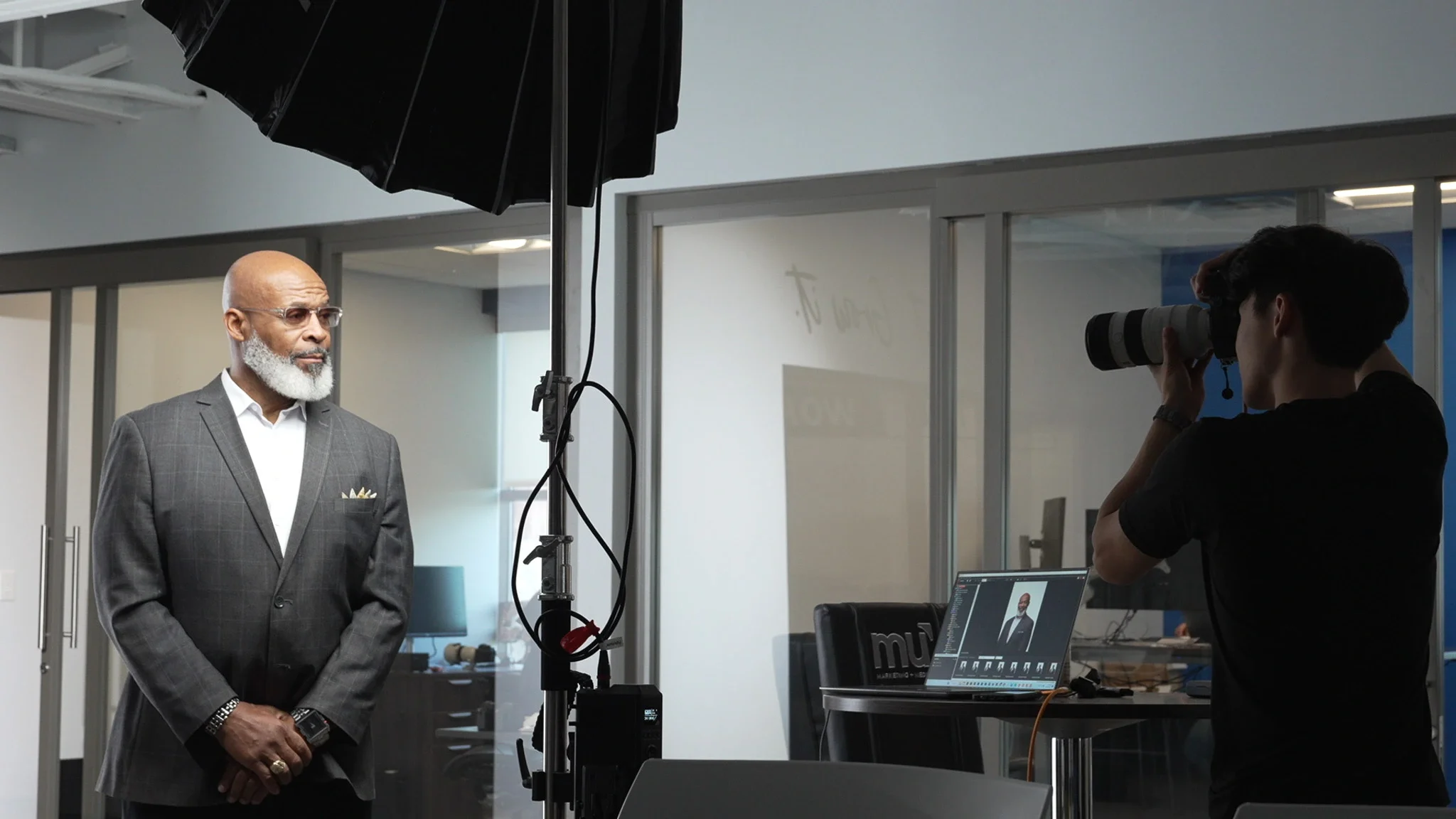 A man with a gray beard and glasses wearing a gray suit and white shirt posing for a photo in a modern office setting. A photographer is taking his picture with a camera, and photography equipment, including a light and a laptop, is visible.