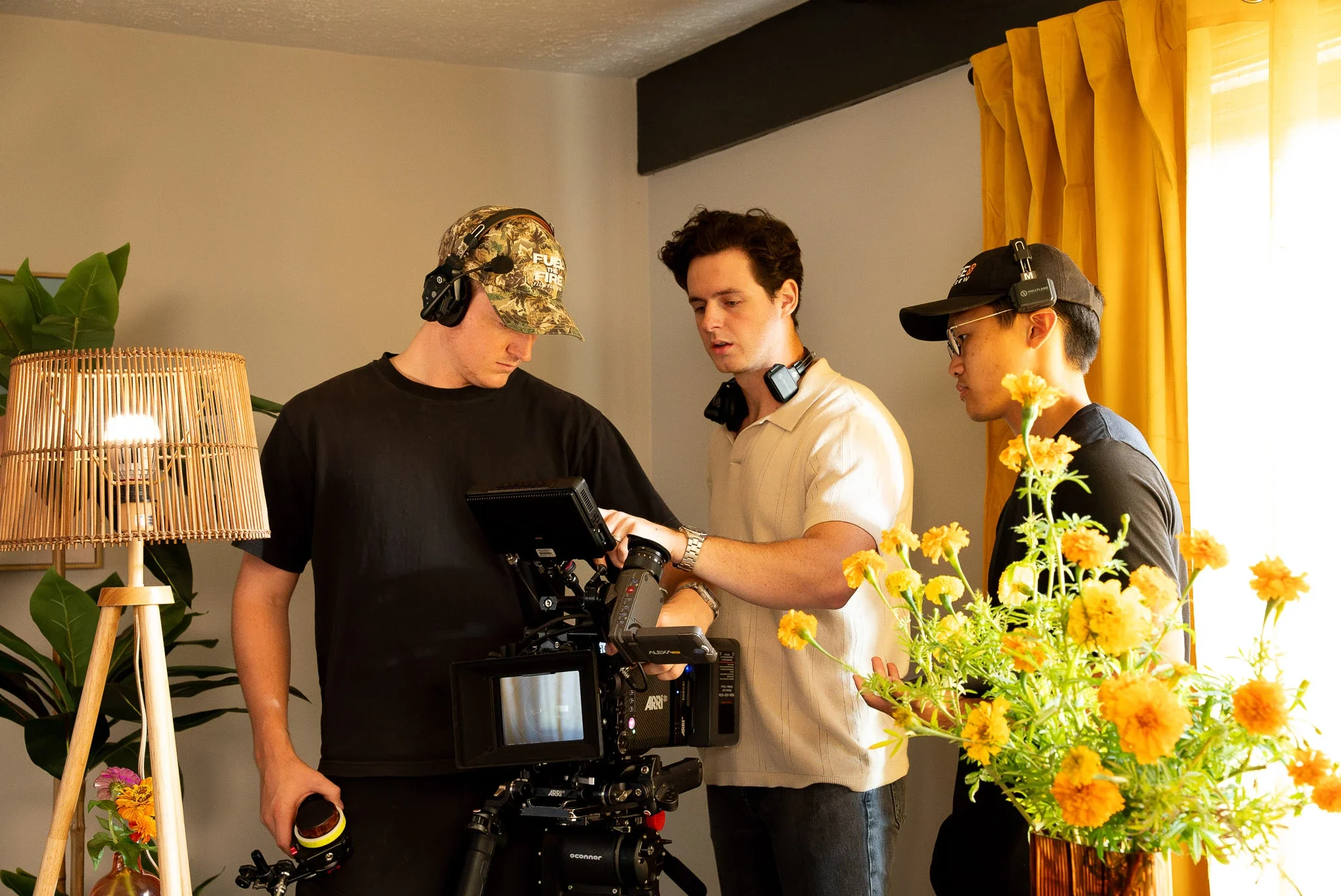 Three young men working together on a film or video project indoors, with a professional camera, headphones, and a bright yellow flower arrangement in the foreground.