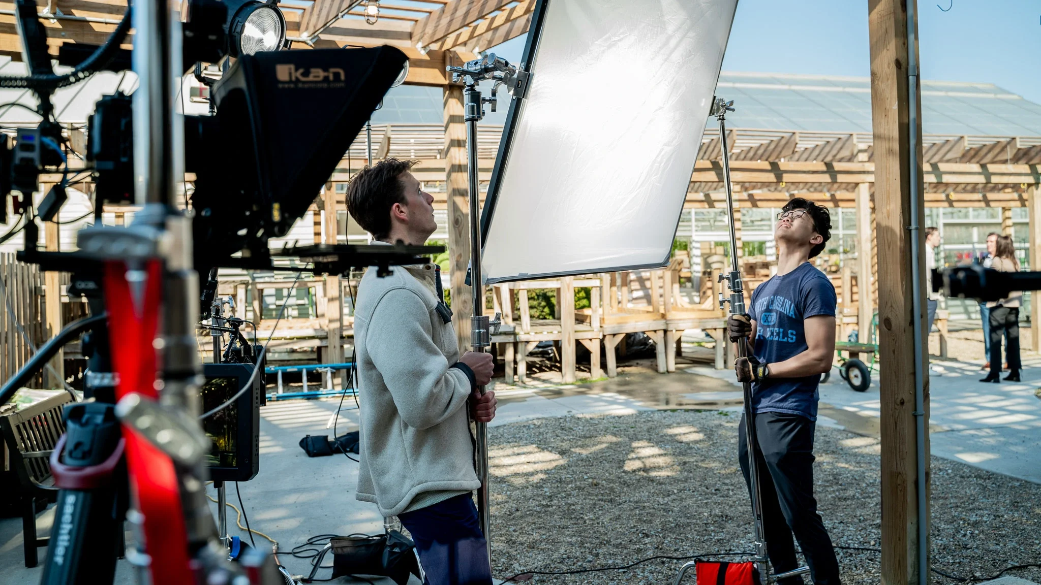 Two young men working on a film shoot outdoors, setting up lighting and camera equipment under a wooden structure.