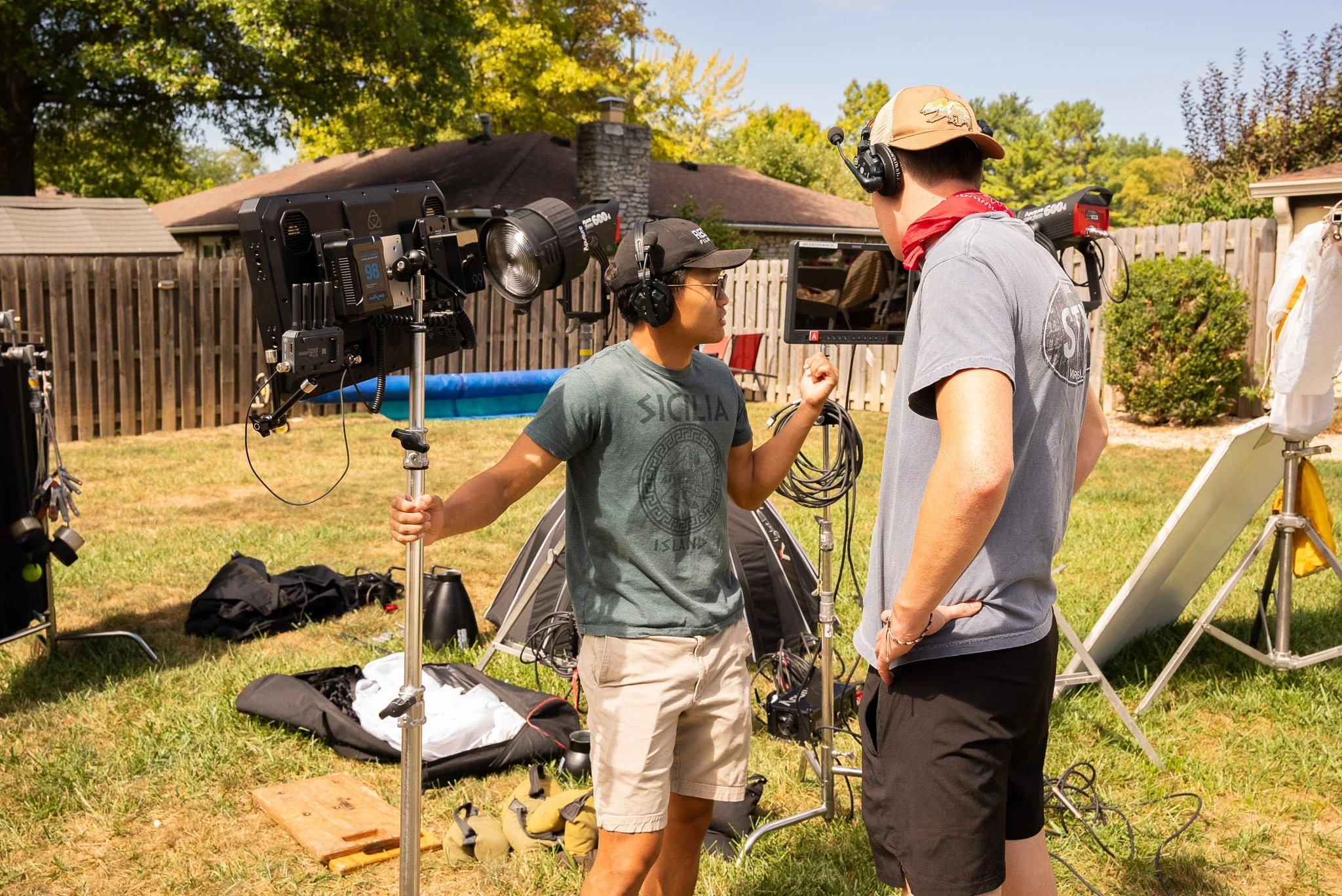 Two crew members on a film set outdoors, with lighting and camera equipment, discussing a scene with a backyard and trees in the background.