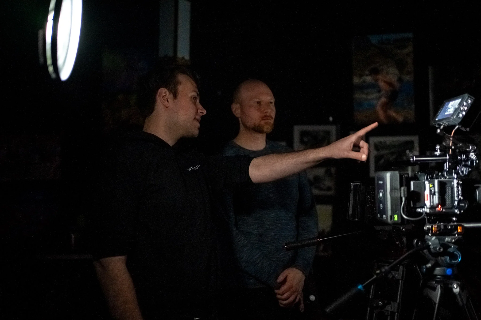 Two men are on a film set, one is pointing at a monitor while the other looks on. The scene is dimly lit.