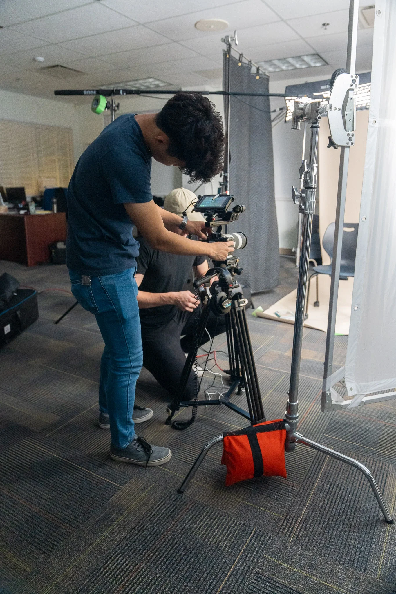 Two videographers set up camera equipment indoors, with one adjusting the camera on a tripod and another standing beside.