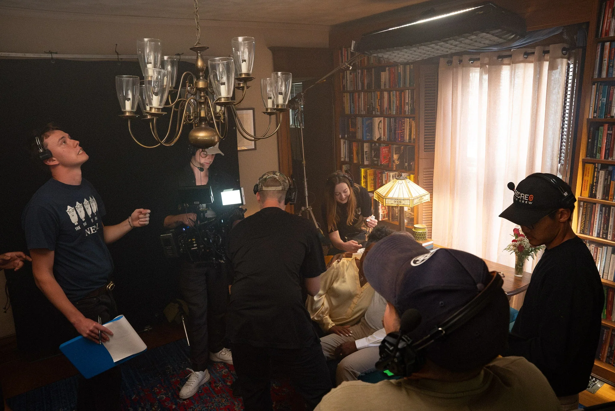 Filming crew working on a movie set in a room with bookshelves, a chandelier, and a window with curtains.
