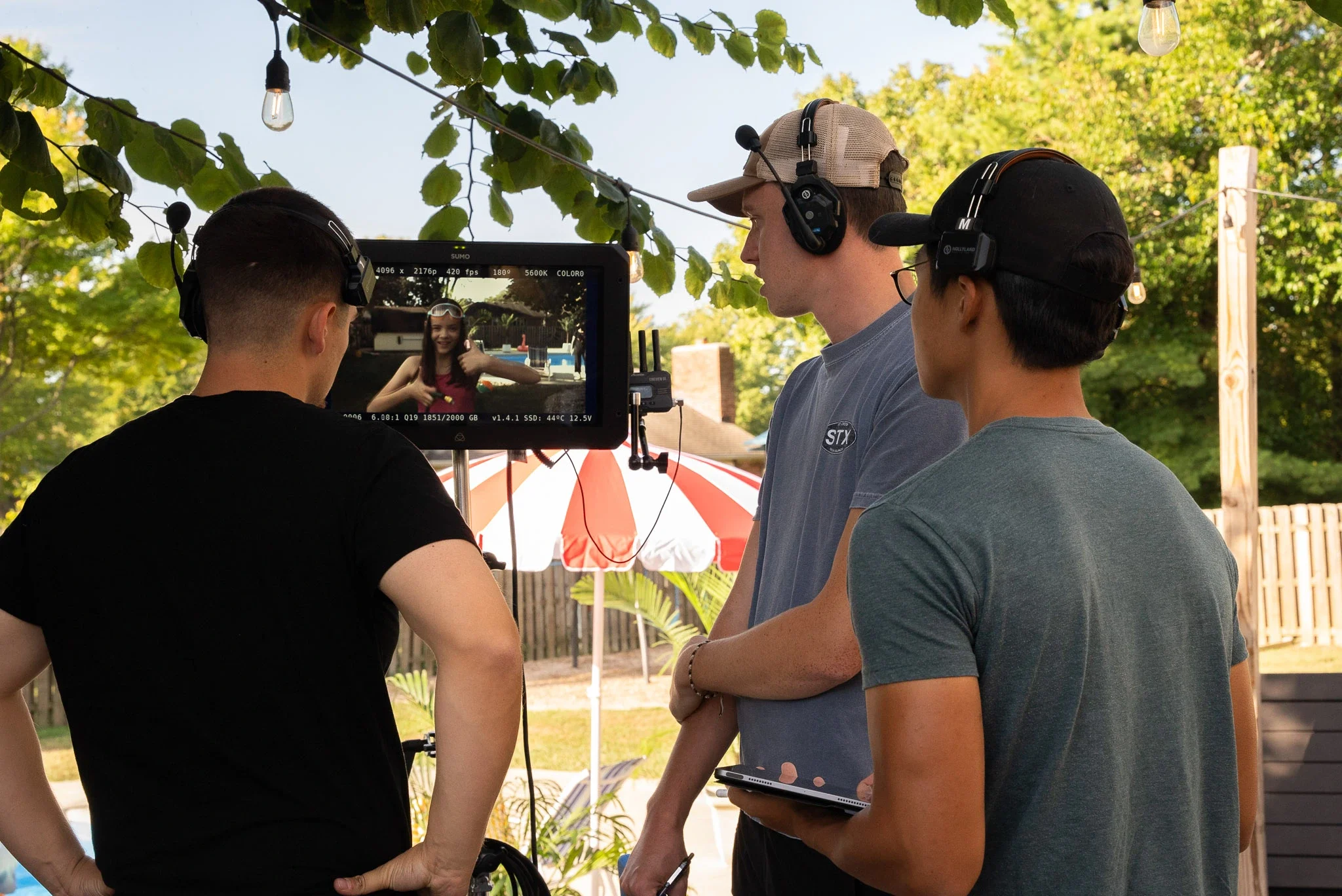 Three people on a film set, with one person operating a camera that has a monitor showing a woman in a red top. The scene takes place outdoors with trees, a wooden fence, and a red and white umbrella in the background.
