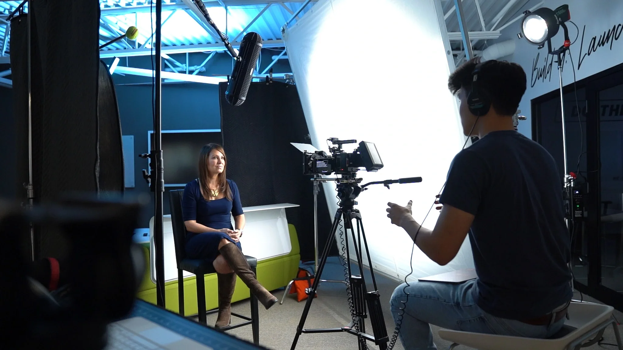 A woman in a navy dress and brown boots sits on a black chair during a video interview in a studio with professional lighting and camera equipment.