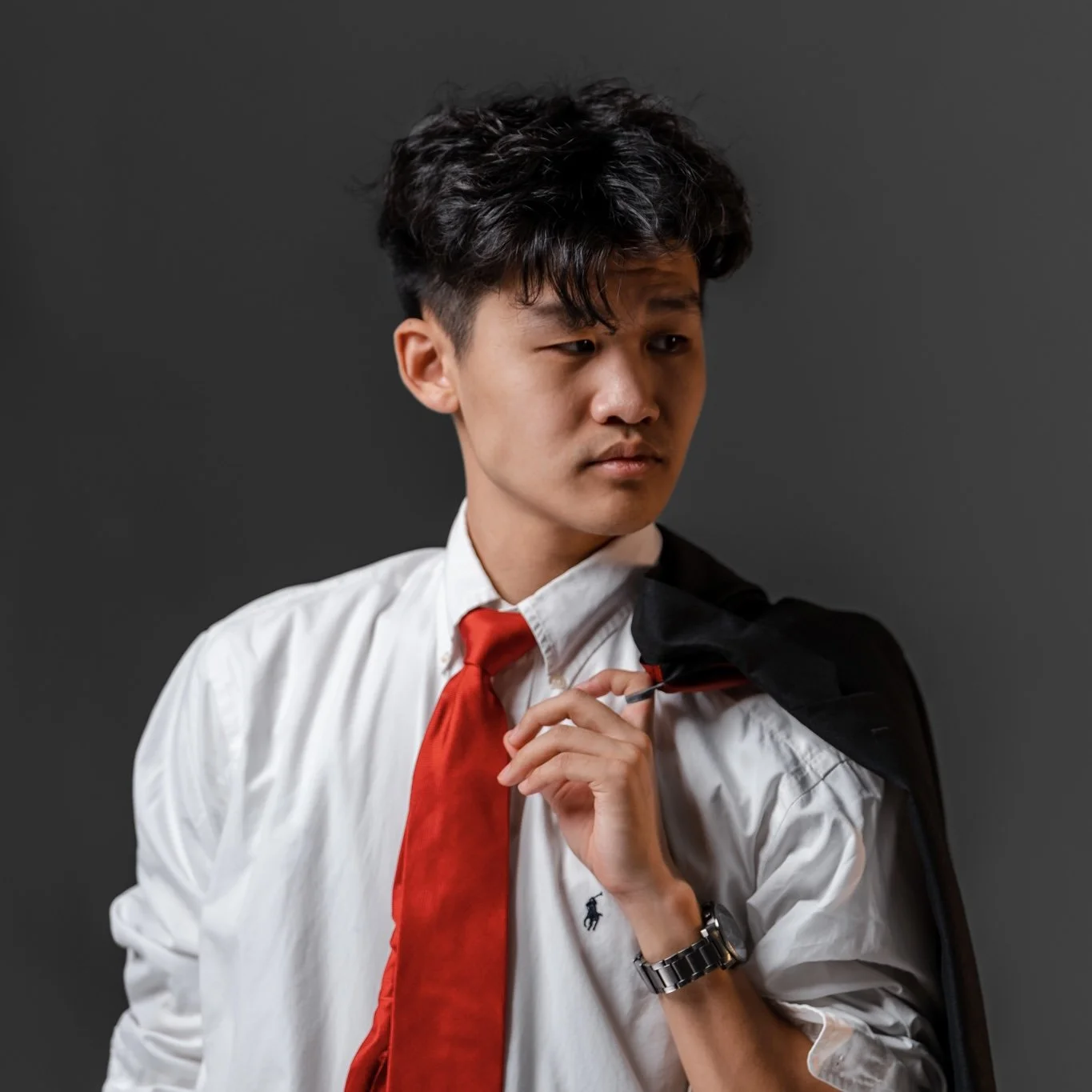 Young man dressed in a white shirt with a red tie, holding a black jacket over his shoulder, standing against a gray background.