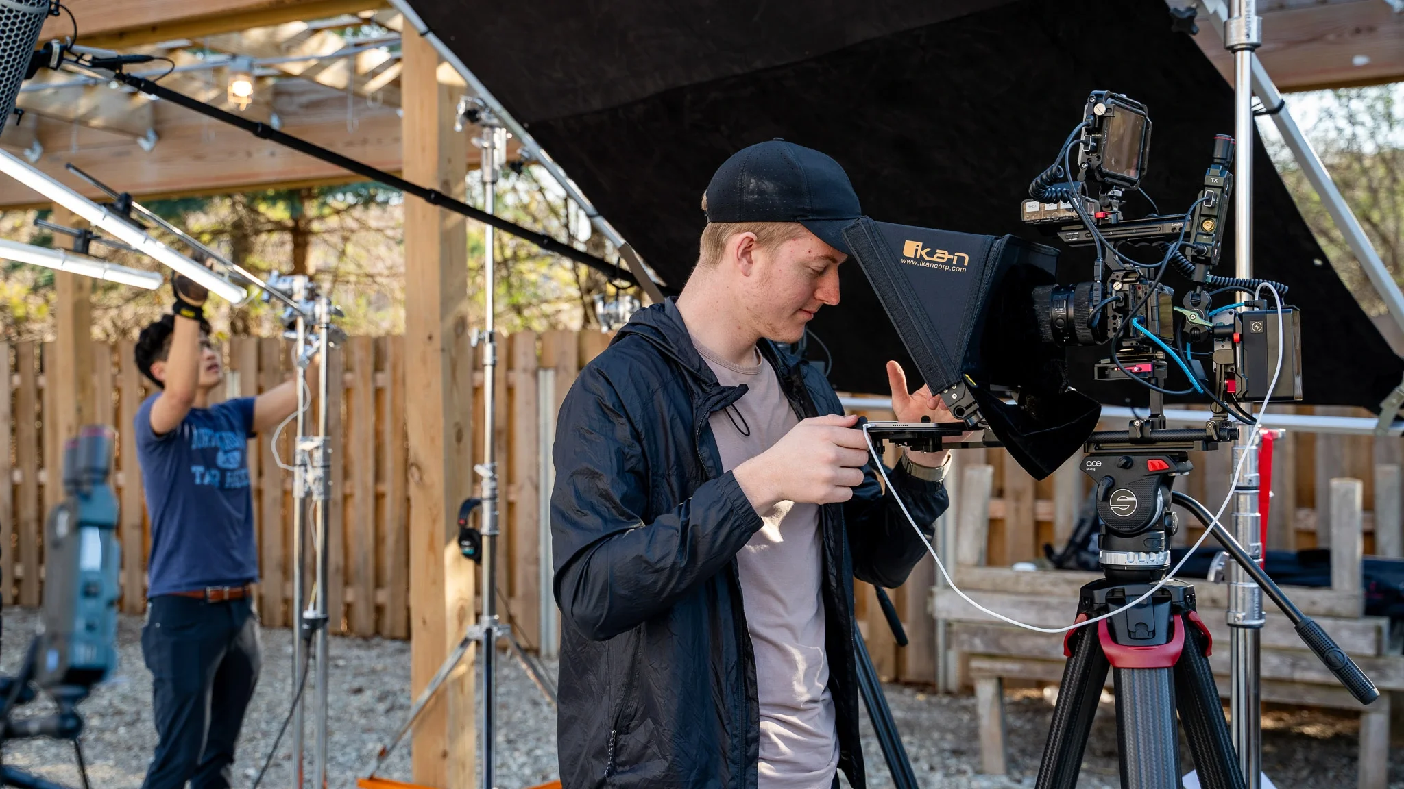 A person operates a large professional camera on a tripod, filming outdoors with a wooden fence in the background. Another person adjusts equipment in the background.