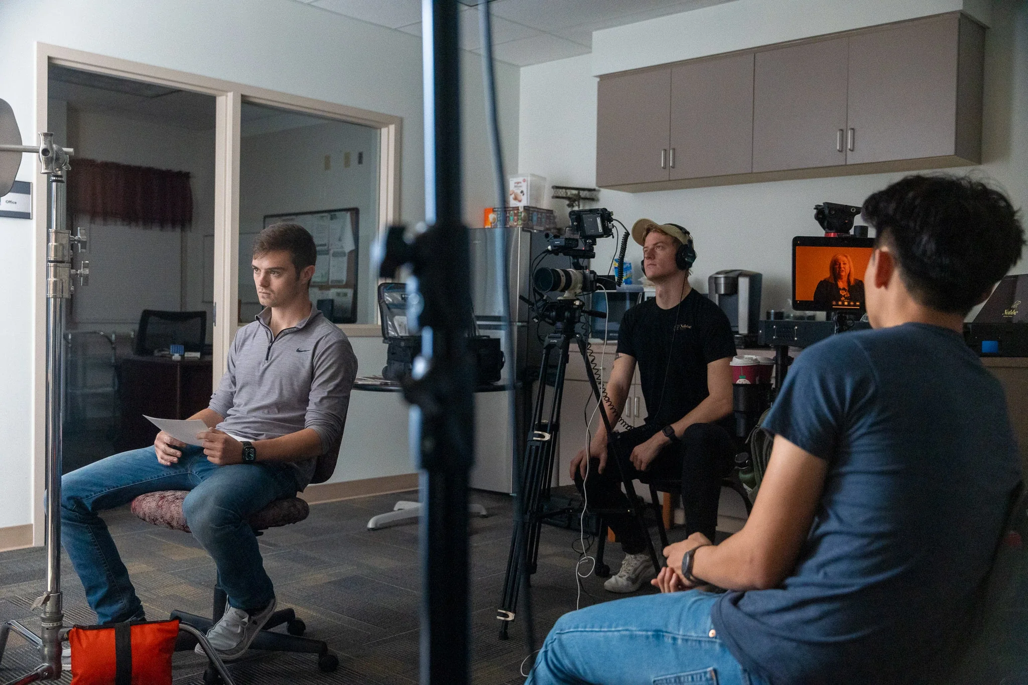 A film crew setting up for an interview in an office, with one man seated holding papers and two crew members operating camera and audio equipment.