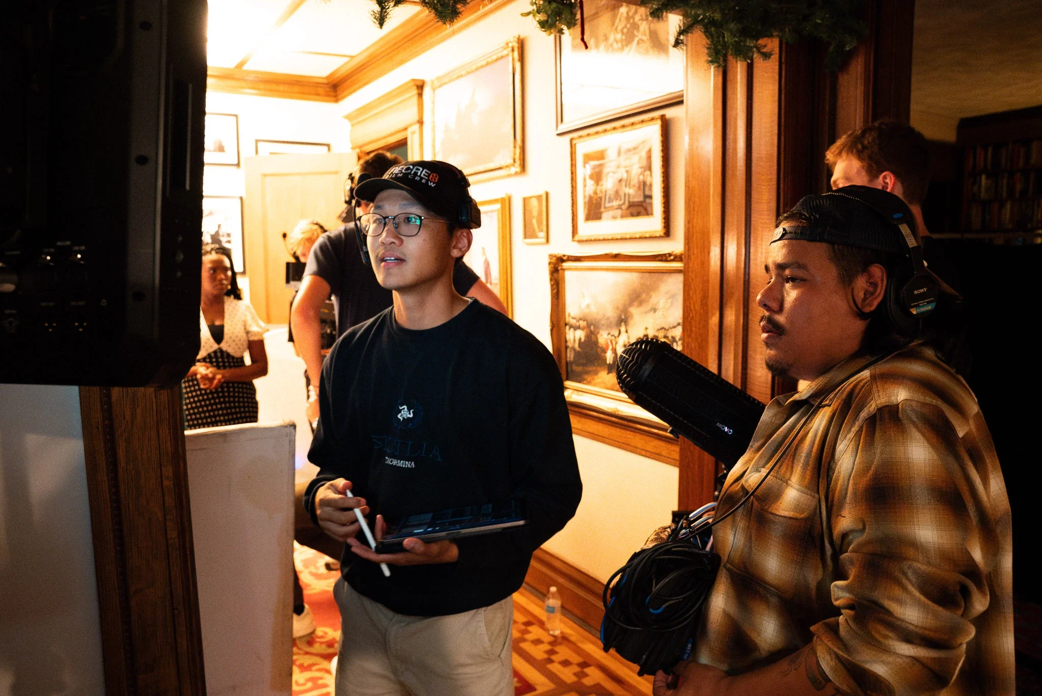 Group of people working on a film or video production inside an ornately decorated room with wooden paneling and framed paintings.