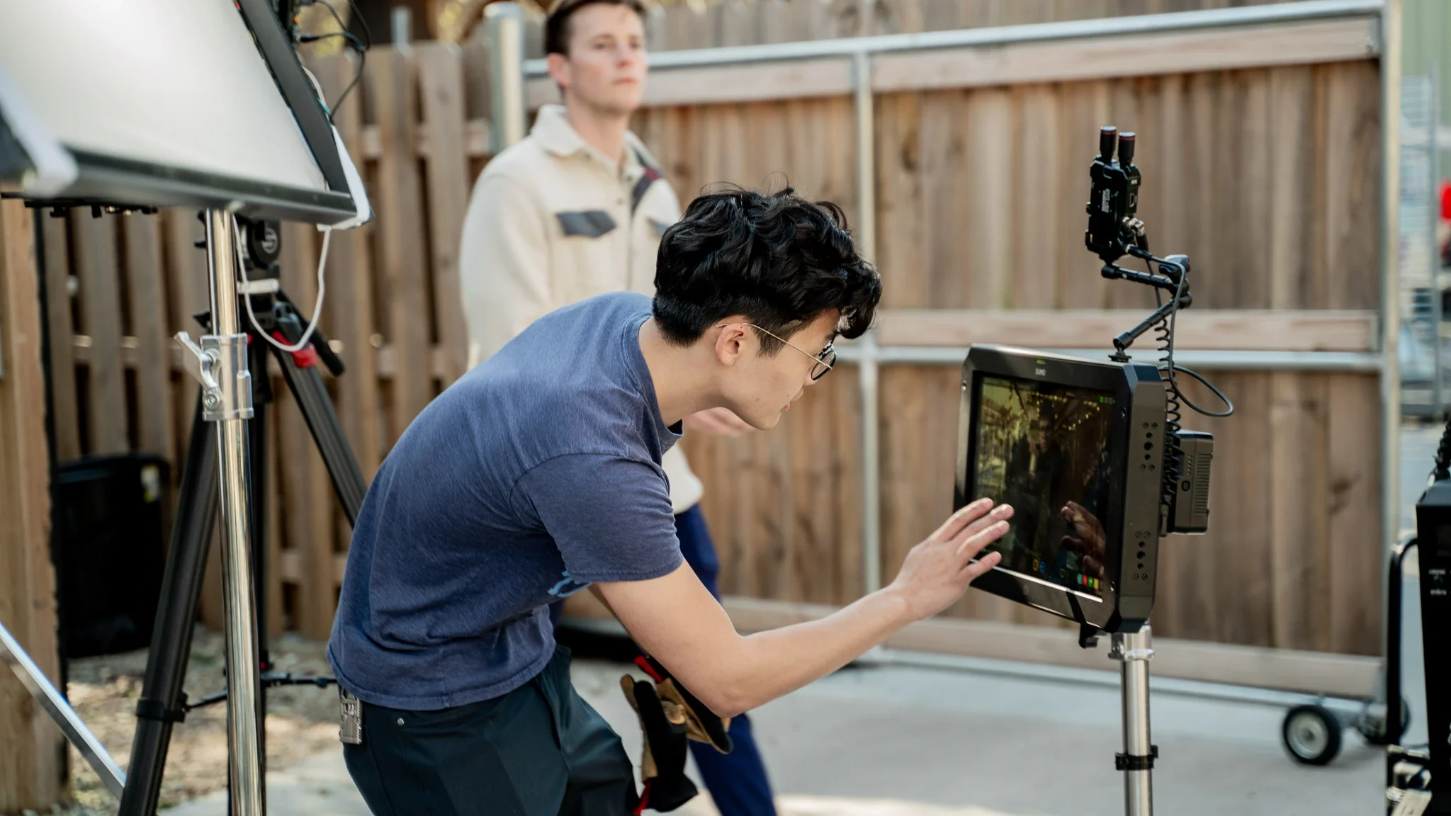 A person in a blue shirt working with a camera monitor on a film set with crew members in the background.