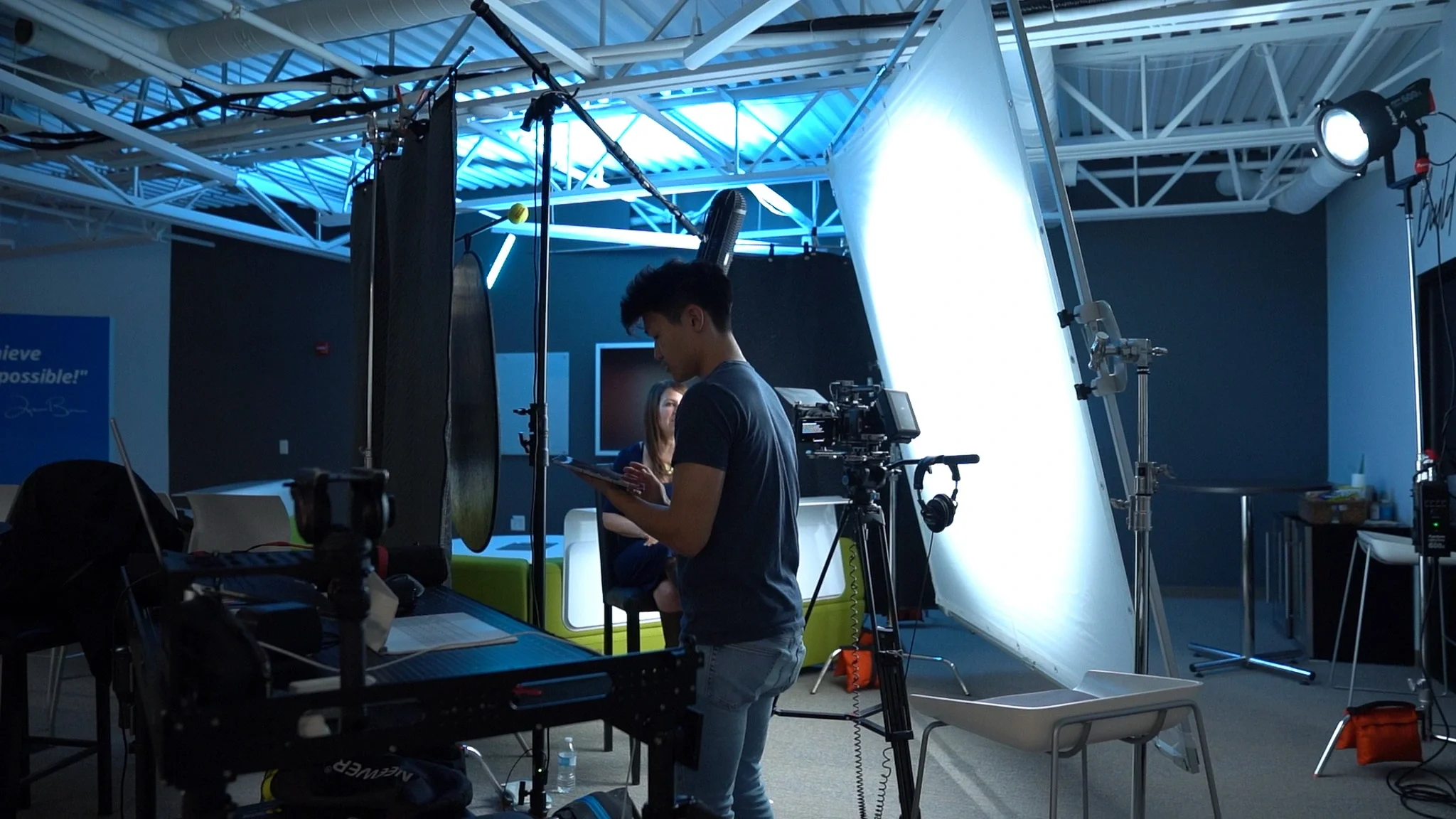 A young man is working in a professional video or photo shoot setup, with lighting equipment, cameras, and a backdrop. A woman is visible in the background on a couch, being filmed or photographed.