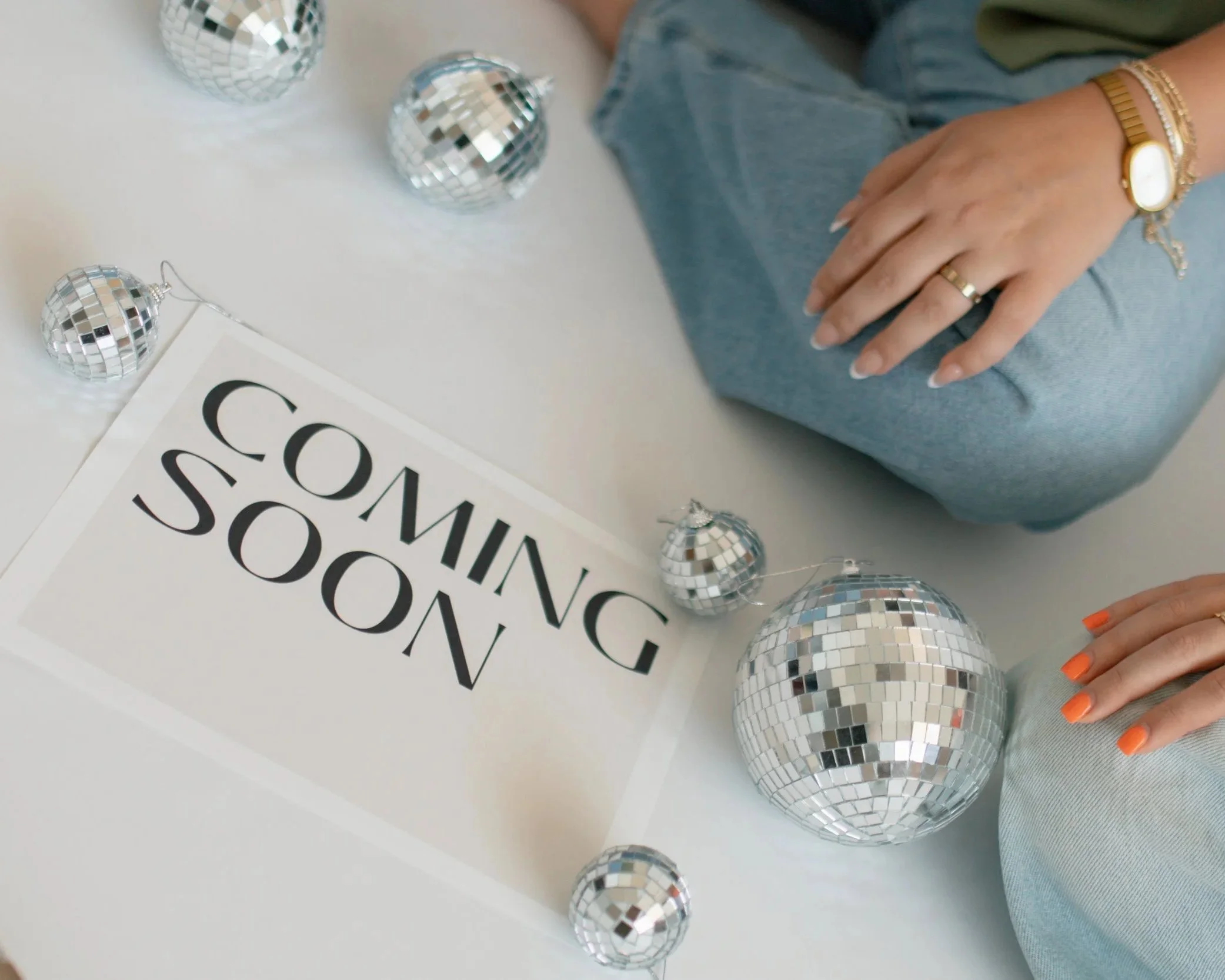 Person sitting next to a sign that says 'Coming Soon' with disco ball ornaments on a white surface.