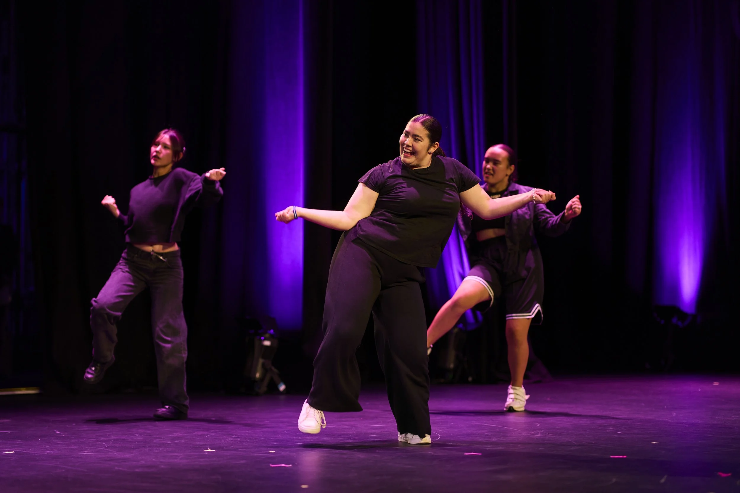 Three women dancing on stage under purple and blue lighting, wearing black athletic outfits.