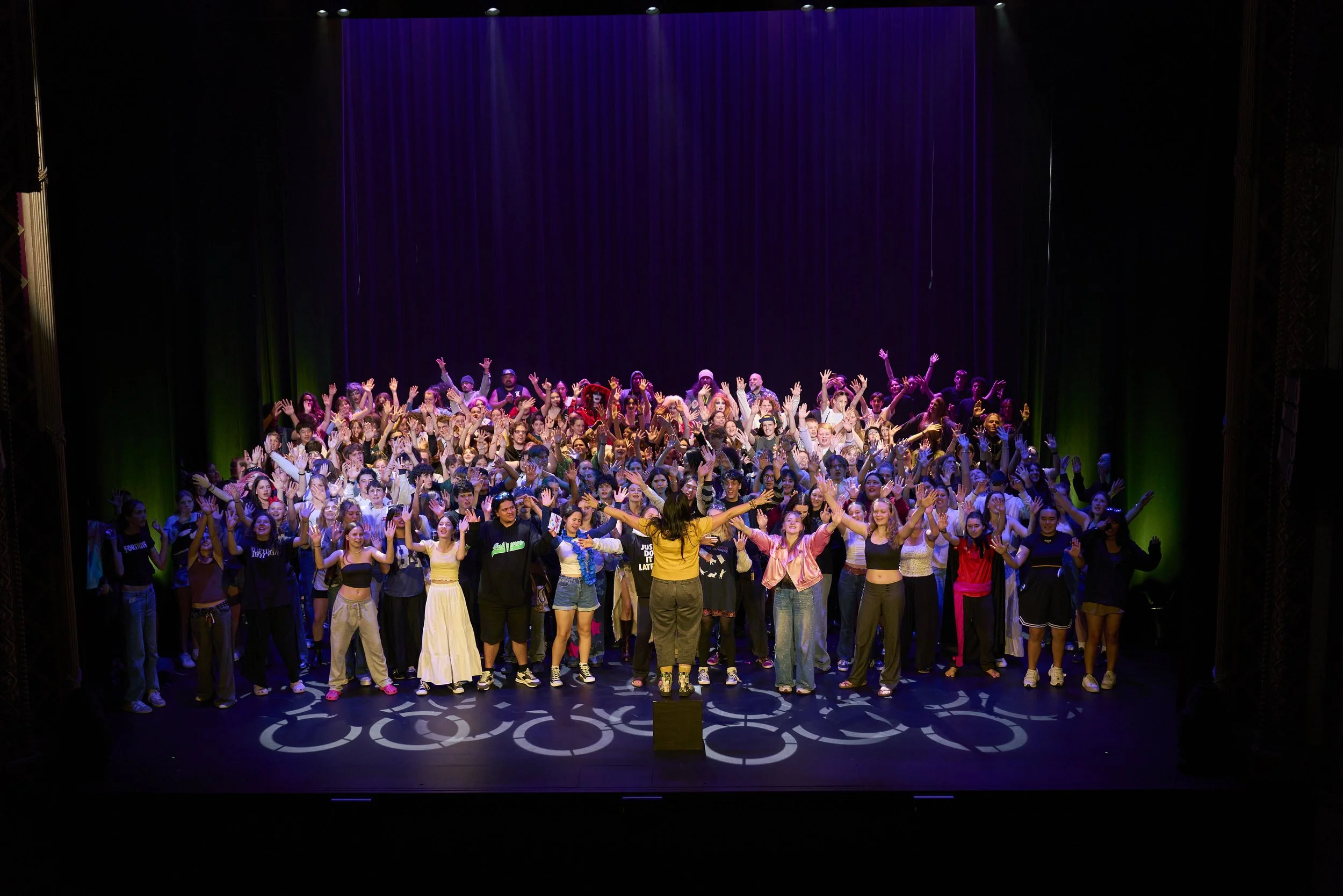 Large group of people performing on stage with a conductor in front, with lines of light projecting bicycle symbols on the floor and purple curtains in the background.