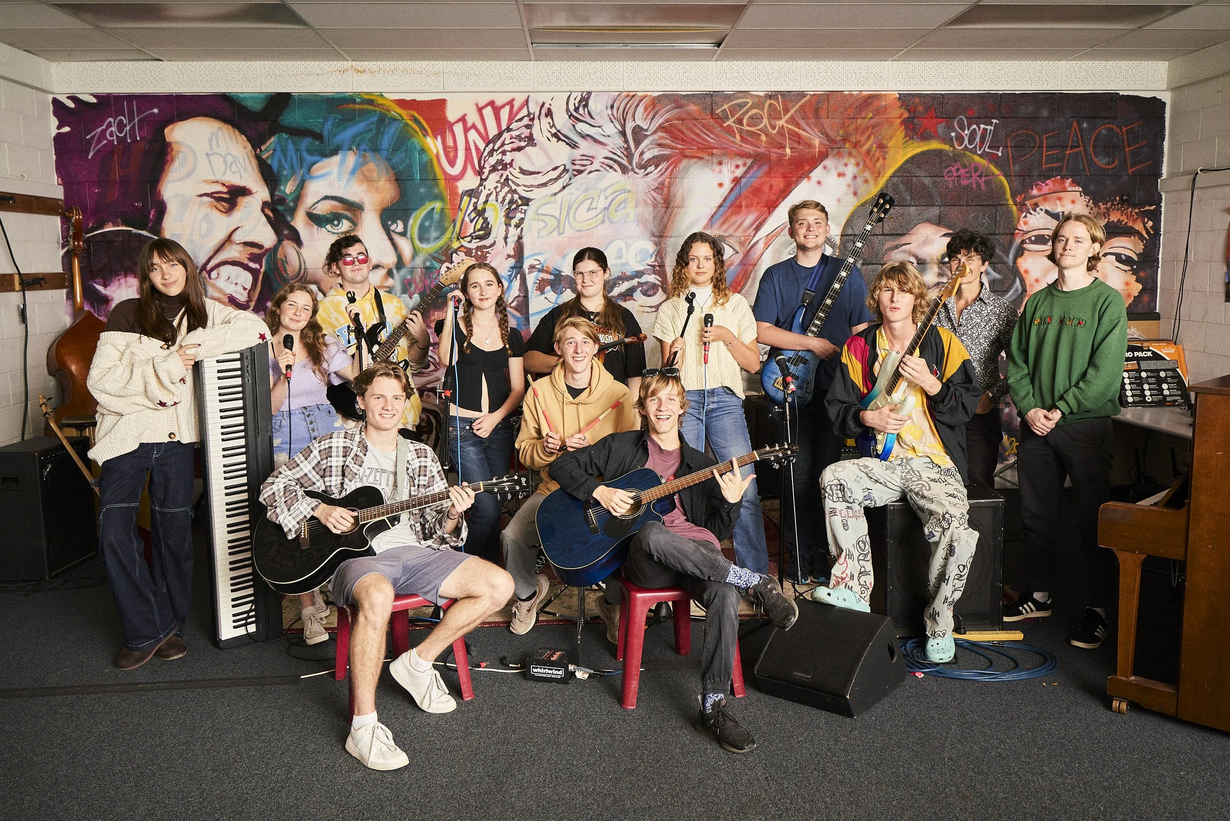 A group of young musicians holding guitars, keyboards, and microphones in a music rehearsal room with a graffiti-style mural of faces and words like "rock" and "peace" on the wall.