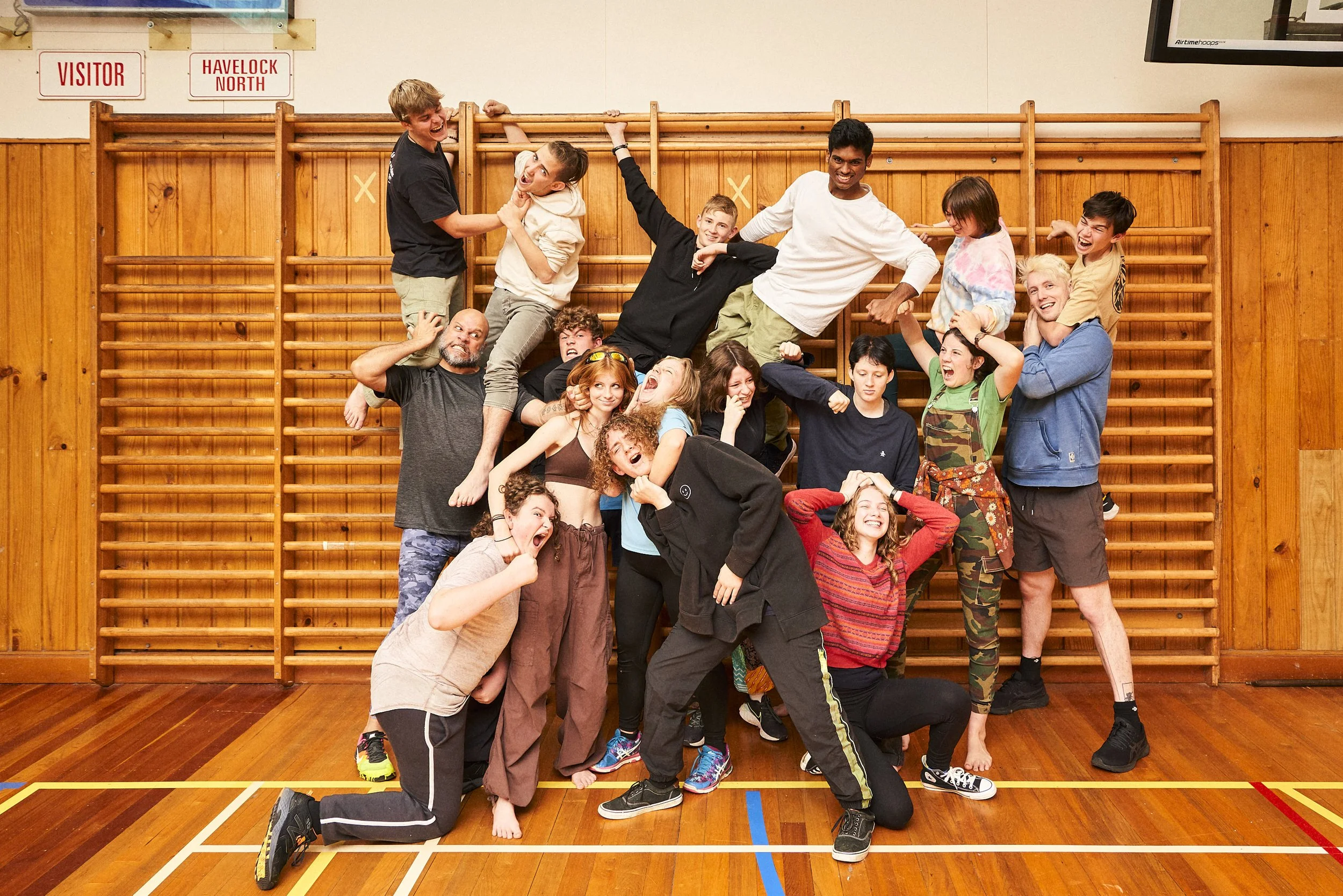 A group of people in a gymnasium posing and playing, with some climbing on a wooden wall and others standing or sitting in front, all smiling and having fun.