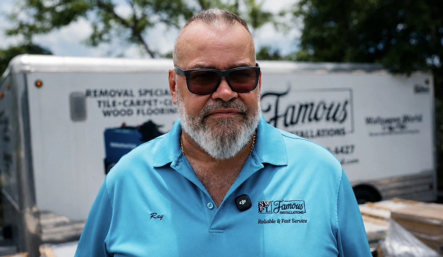 A middle-aged man with a gray beard and sunglasses wearing a light blue polo shirt with embroidered text and a badge, standing outdoors in front of a moving truck.