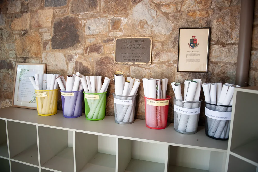 Coloured tubs of rolled brochures and posters on a shelf against the stone wall.